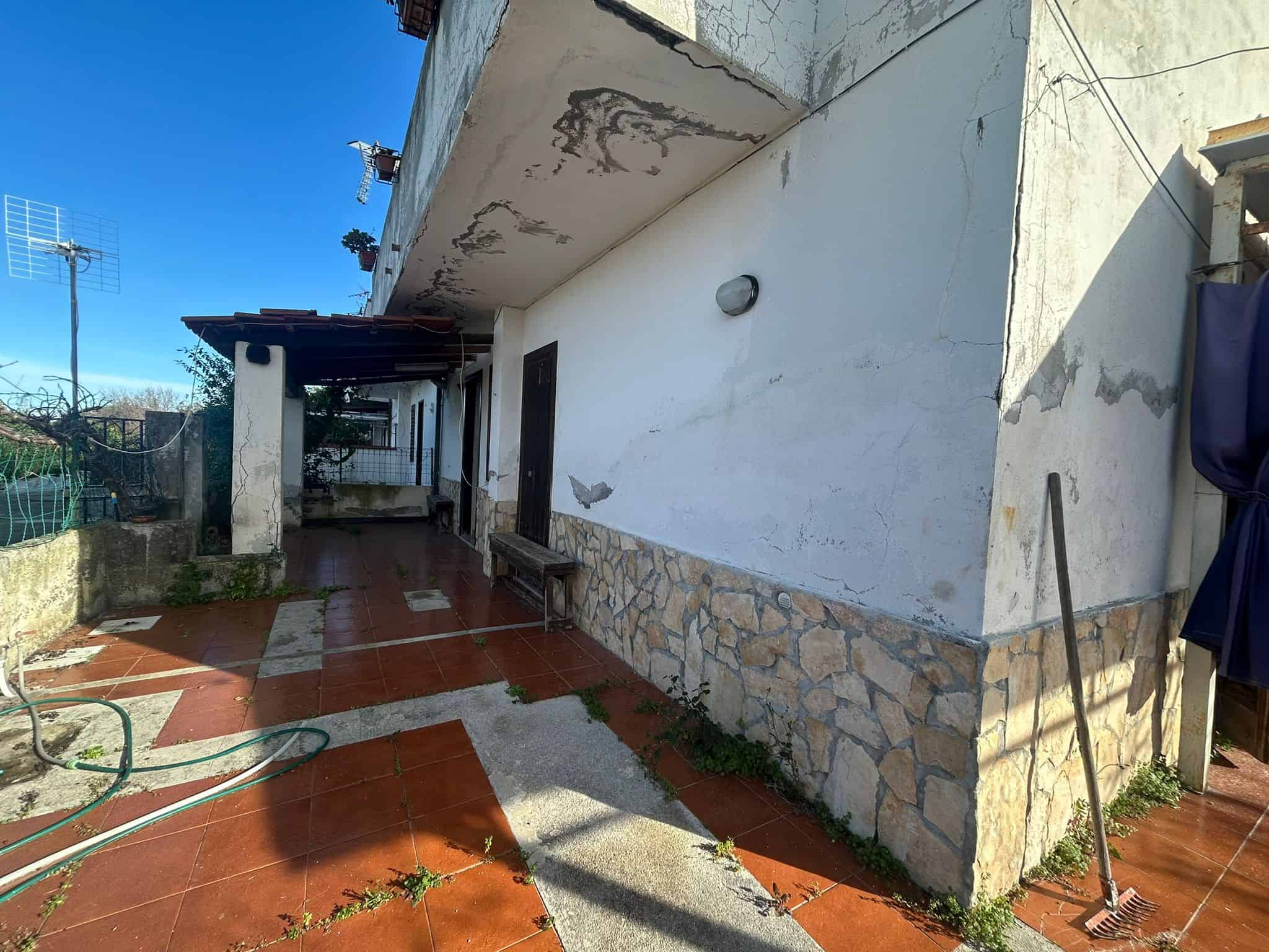 Tetto e facciata di una casa in cattive condizioni con muri danneggiati e pittura scrostata, patio con piastrelle rosse e piante rampicanti, strumenti da giardino, ambiente esterno rustico e trasandato in cerca di ristrutturazione.