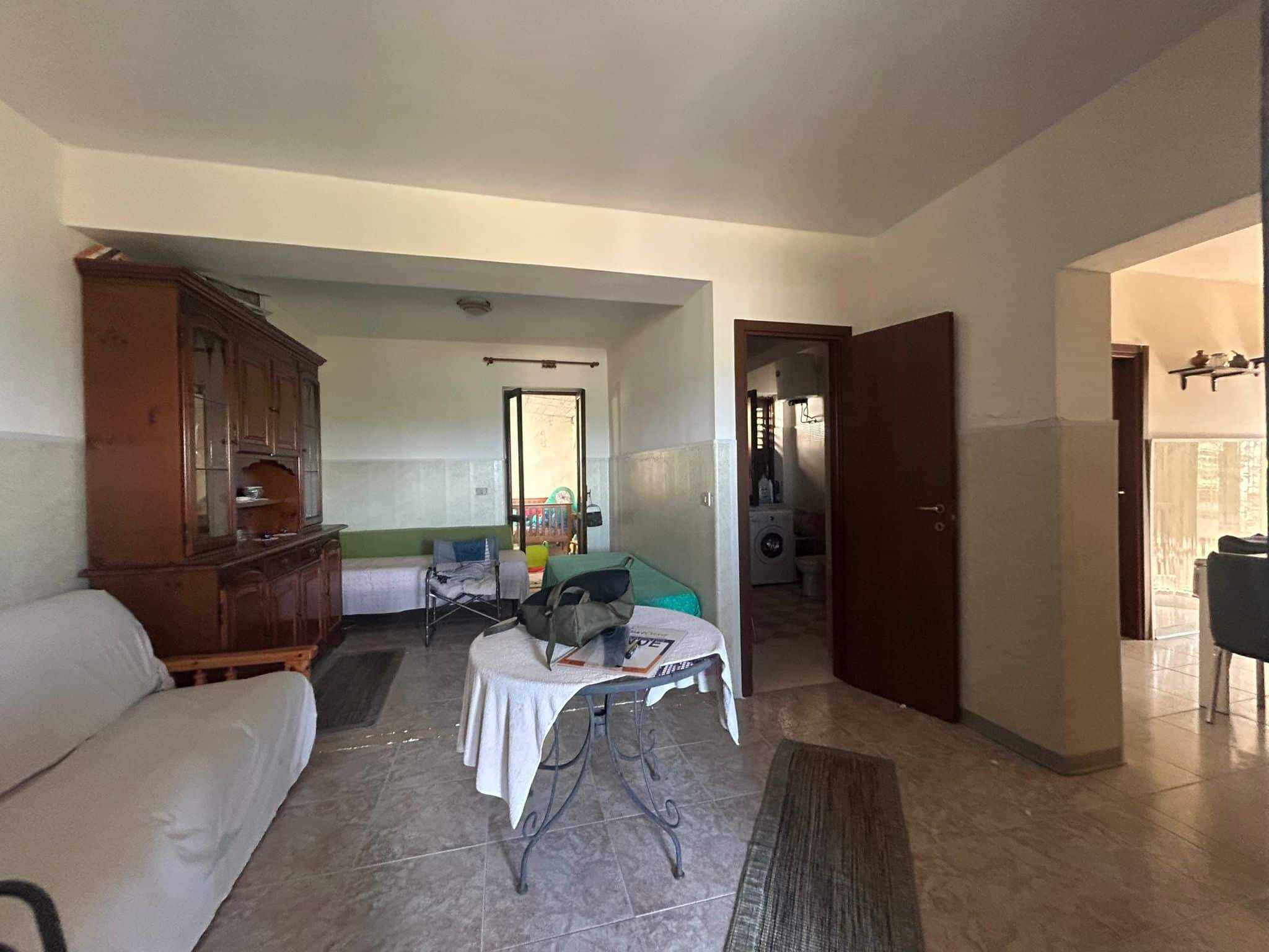 Spazio abitativo con divano bianco, mobili in legno, porta verso balcone, area lavanderia e accesso alle altre stanze. Atmosfera familiare e accogliente, ideale per vivere in tranquillità e comodità.