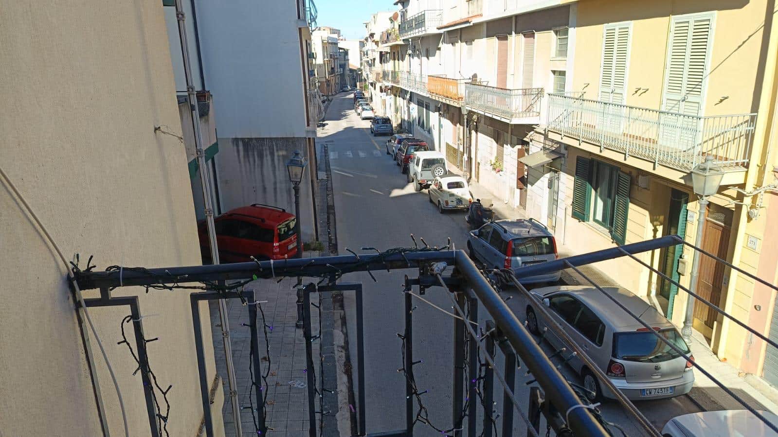 Vista di una strada urbana con veicoli parcheggiati, edifici residenziali con balconi e finestre, lampioni storici, atmosfera luminosa e soleggiata, ideale per il mercato immobiliare e la ricerca di appartamenti a NextCasa.