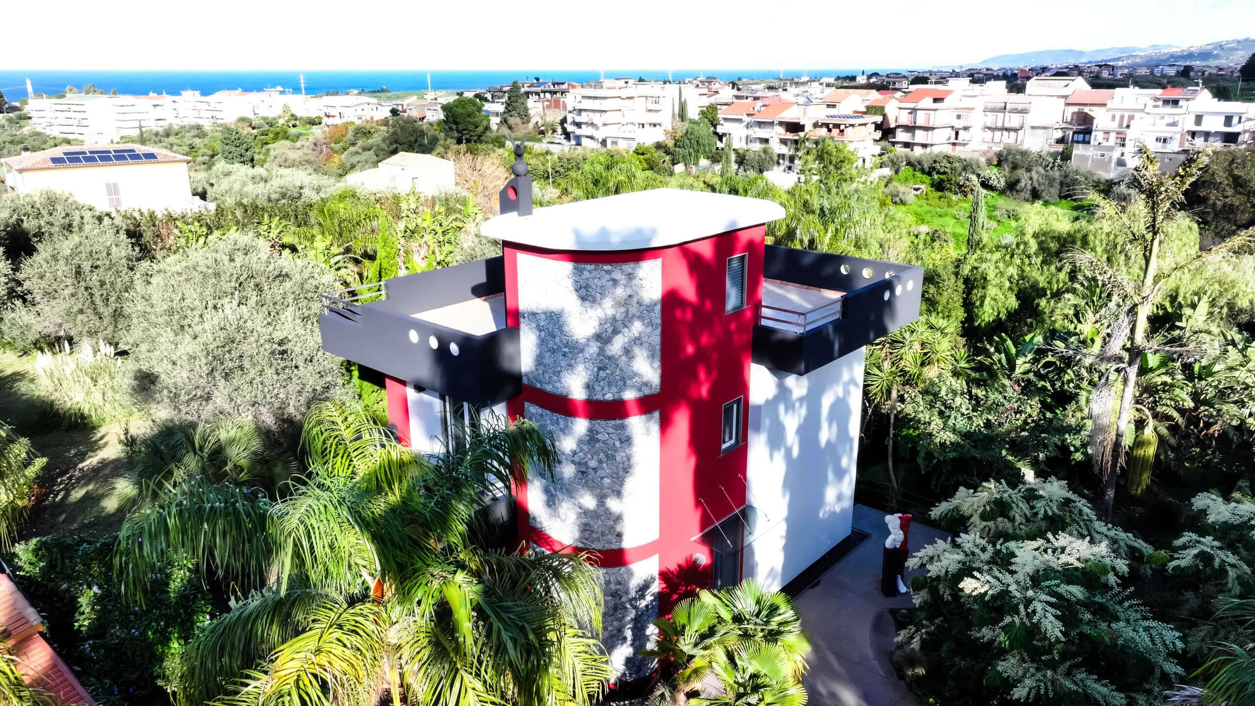 Vista aerea di una casa moderna con dettagli artistici e testo NextCasa, immersa in un giardino lussureggiante con piante tropicali, con vista sul mare e sul paesaggio urbano circostante in stile contemporaneo.