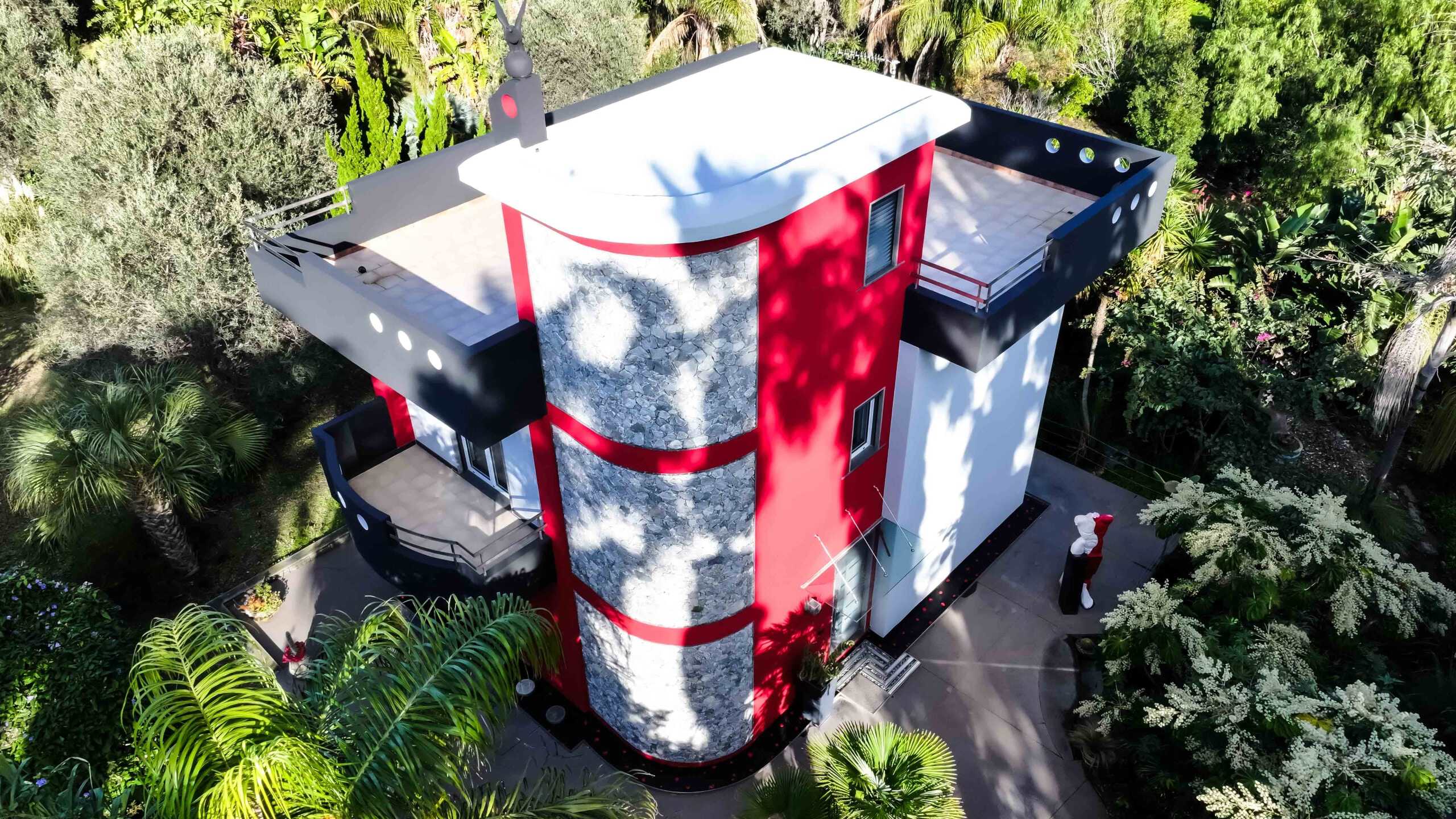 Elegante villa moderna con facciata rossa e dettagli in pietra, circondata da vegetazione tropicale, terrazza panoramica e design innovativo, ideale per residenza di lusso e relax all'aperto.
