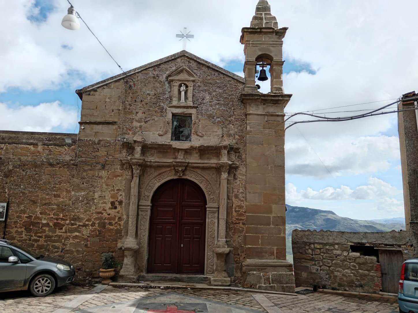 Portale in pietra della chiesa storica in campagna, con campanile e dettagli architettonici antichi, in un paesaggio rurale con montagne sullo sfondo.