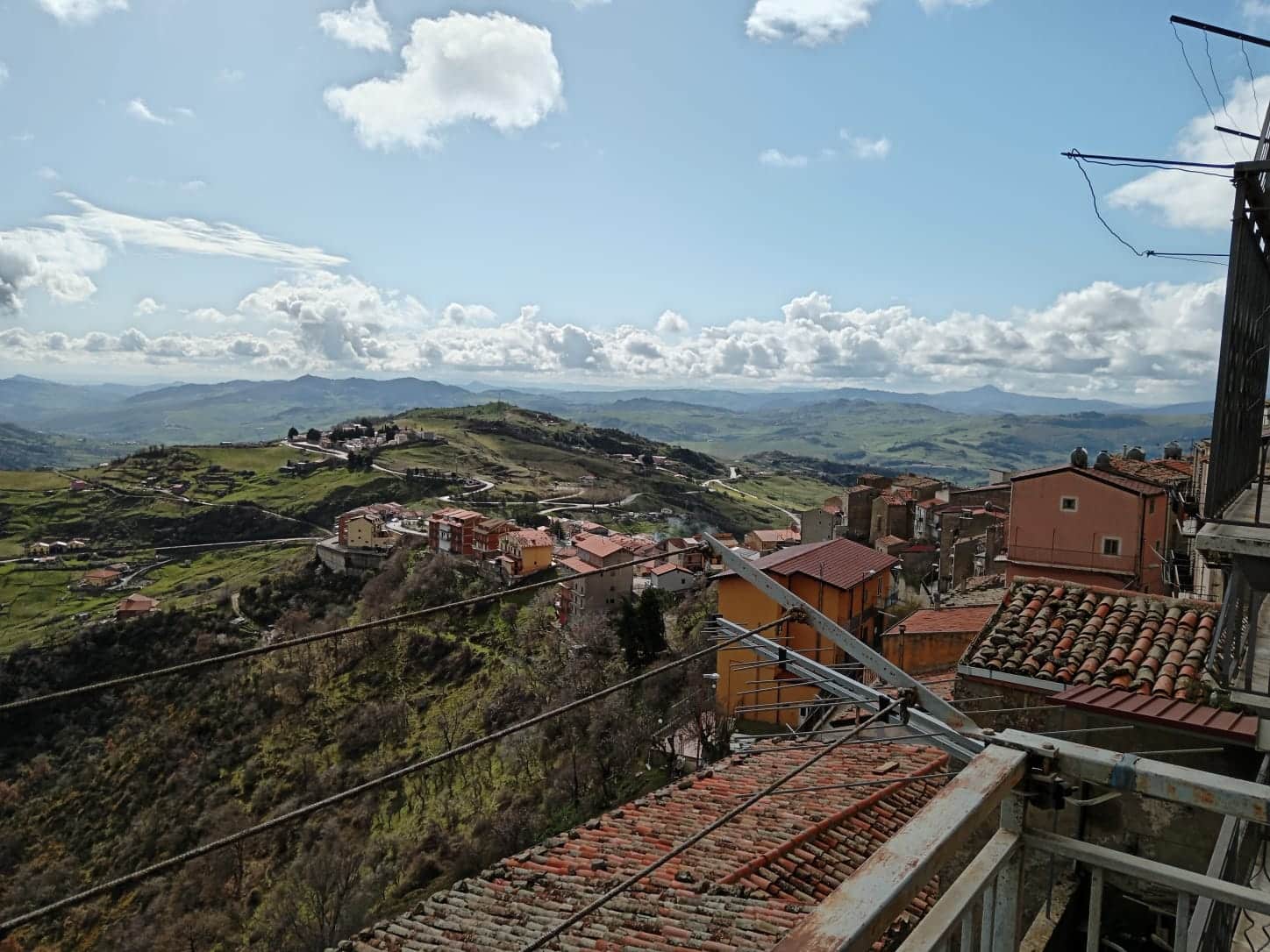 Paesaggio montano con case colorate e terreni verdi, vista panoramica sulle colline e cielo nuvoloso, scena da balcone o terrazza di un'abitazione in un villaggio rurale italiano.
