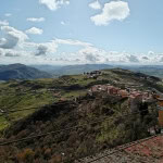 Paesaggio collinare siciliano con case tradizionali, strade tortuose e verdeggianti terreni agricoli, vista panoramica sulle colline e montagne sotto un cielo nuvoloso, ideale per promuovere immobili e vacanze in Sicilia.