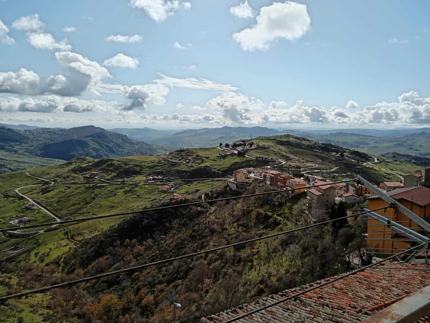 Paesaggio collinare siciliano con case tradizionali, strade tortuose e verdeggianti terreni agricoli, vista panoramica sulle colline e montagne sotto un cielo nuvoloso, ideale per promuovere immobili e vacanze in Sicilia.