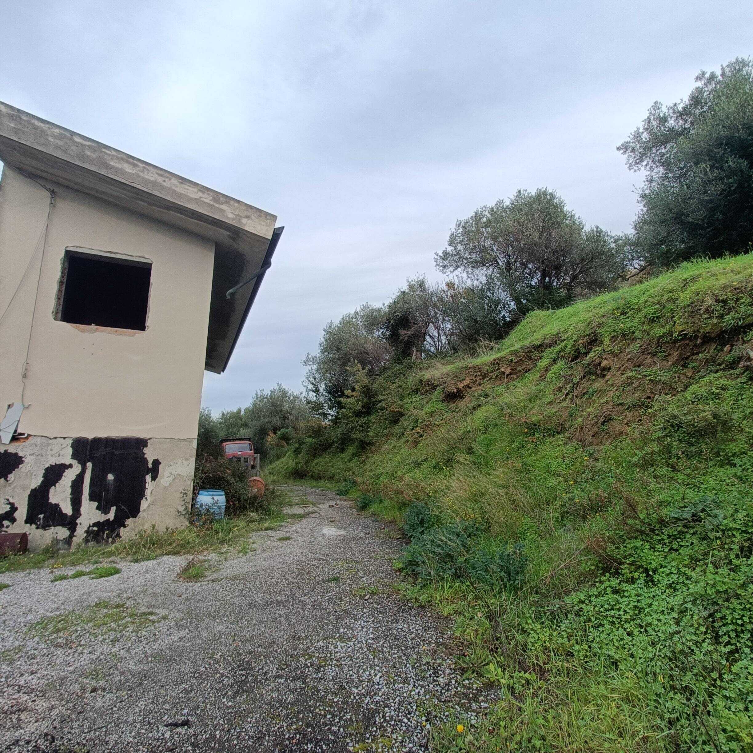 Vecchio immobile in stato di abbandono con pareti bianche e finestra senza vetri, situato su un terreno con vegetazione e un sentiero sterrato, in contesto rurale e naturale.