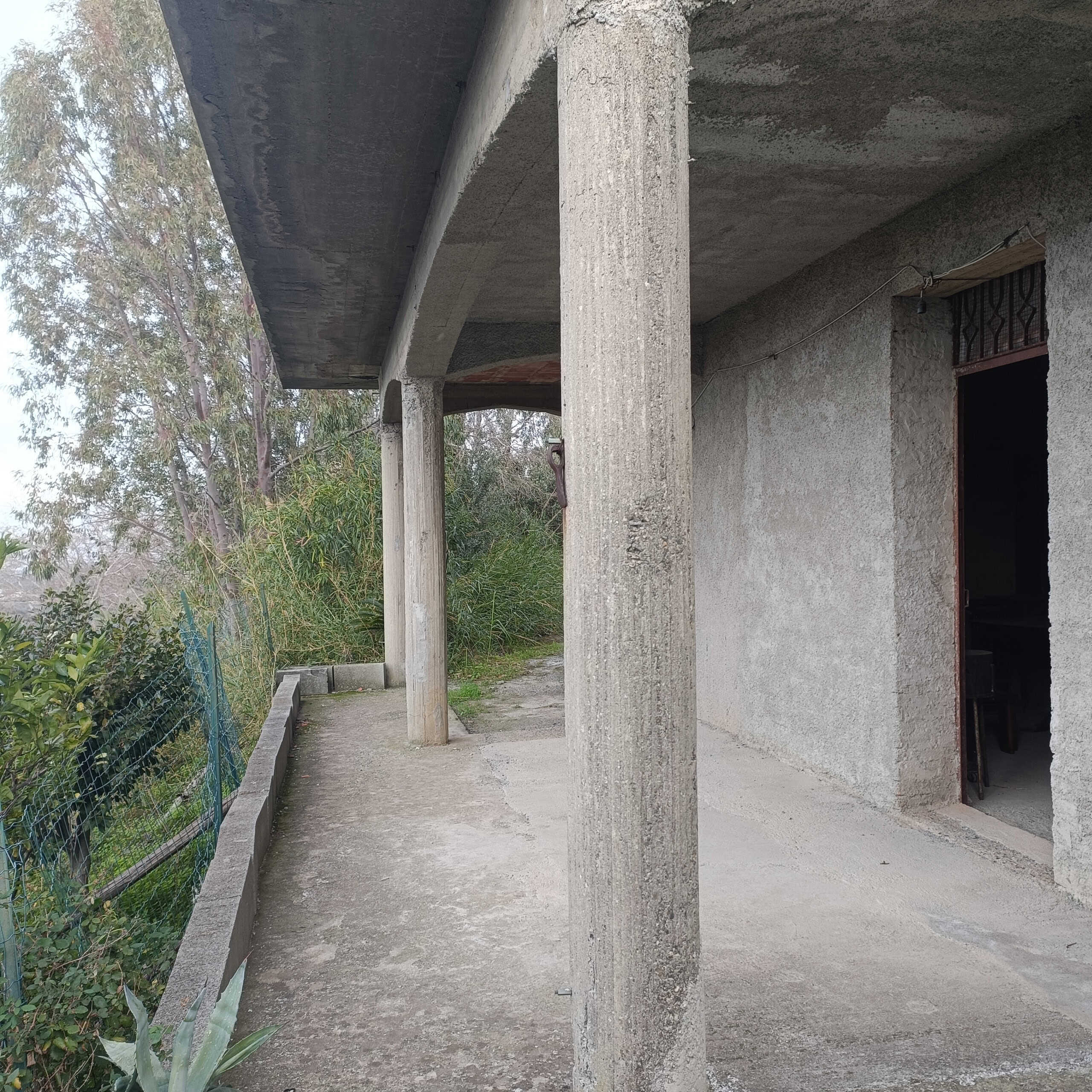 Fuga di casa in costruzione, verifica esterna con colonne in cemento e pareti in calcestruzzo, area aperta e giardino verde circostante, intervento edilizio in fase di completamento.