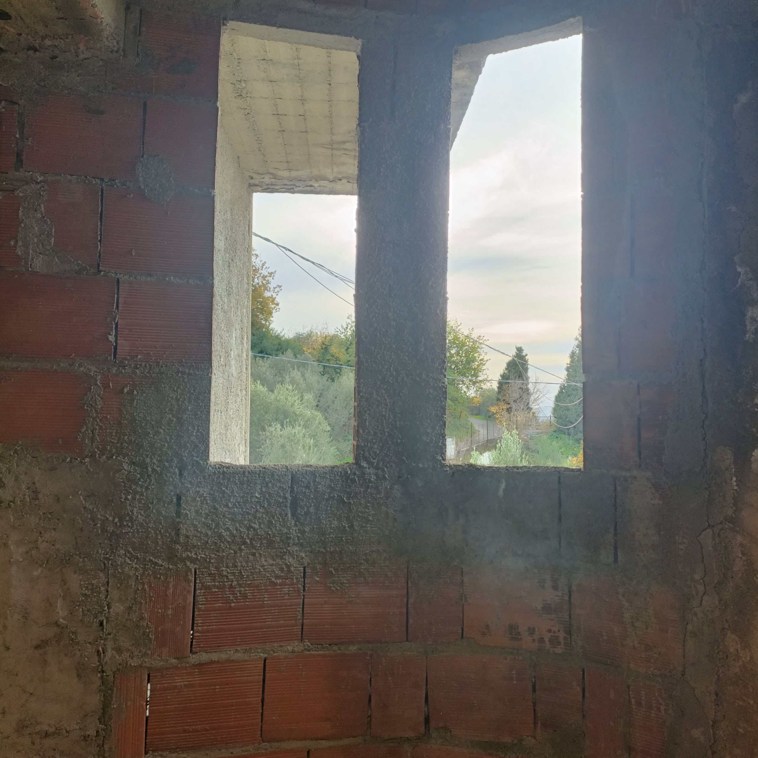 Finestra in costruzione di una casa con pareti in mattoni rossi e cemento grezzo, vista panoramica sulla natura all'aperto, illuminata dalla luce naturale del giorno.