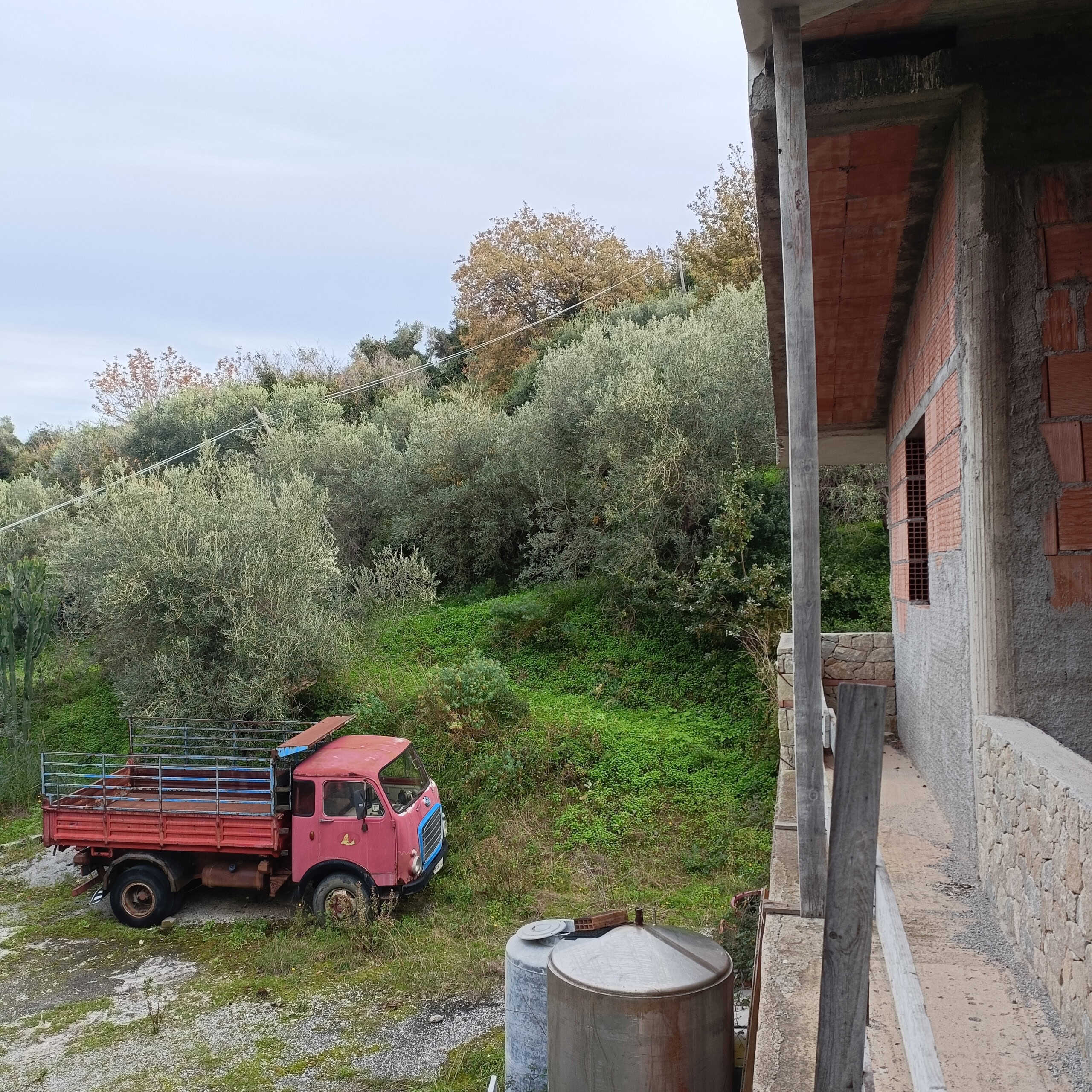 Camion rosso abbandonato nei dintorni di una casa in costruzione, con vegetazione e alberi sullo sfondo, viste dall'angolo di una veranda in fase di realizzazione.