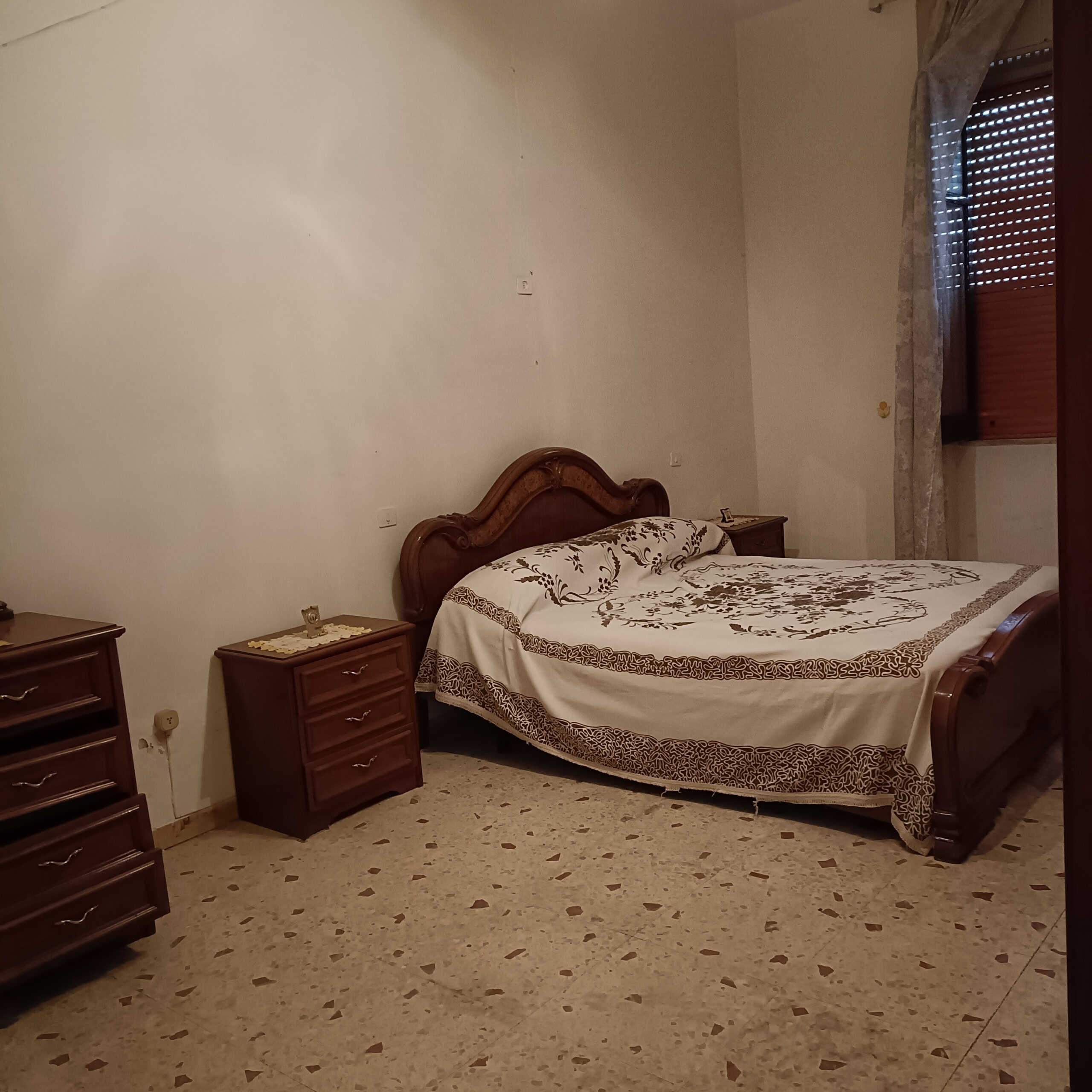 Letto matrimoniale in stile classico con grande testiera in legno, mobili da camera coordinati, e finestra con persiana e tendaggi.