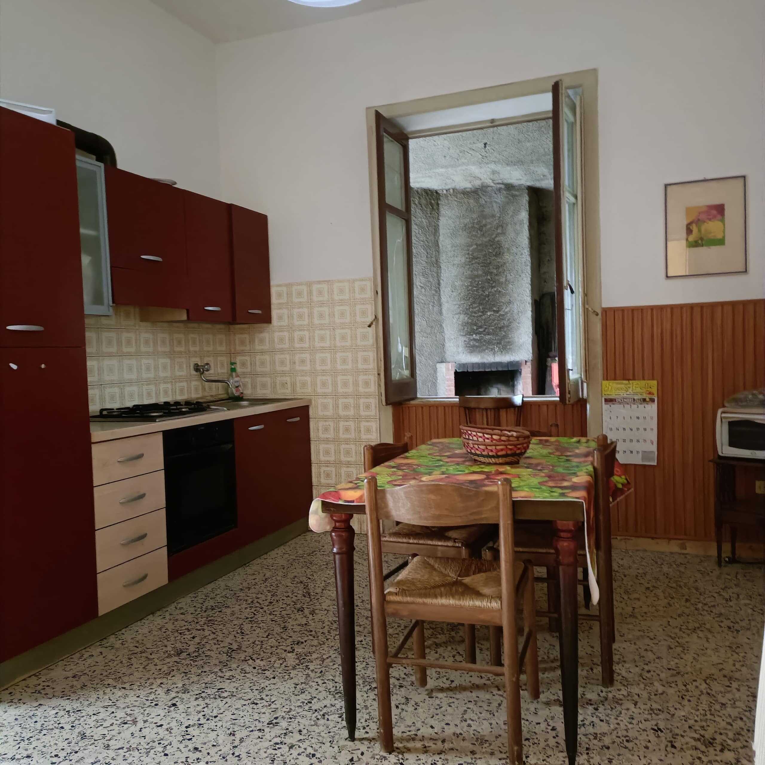 Vecchia cucina rustica con tavolo in legno, sedia e finestra che offre vista sul cortile interno, perfetta per stanze di case vacanza o proprietà storiche in vendita, in stile tradizionale italiano.