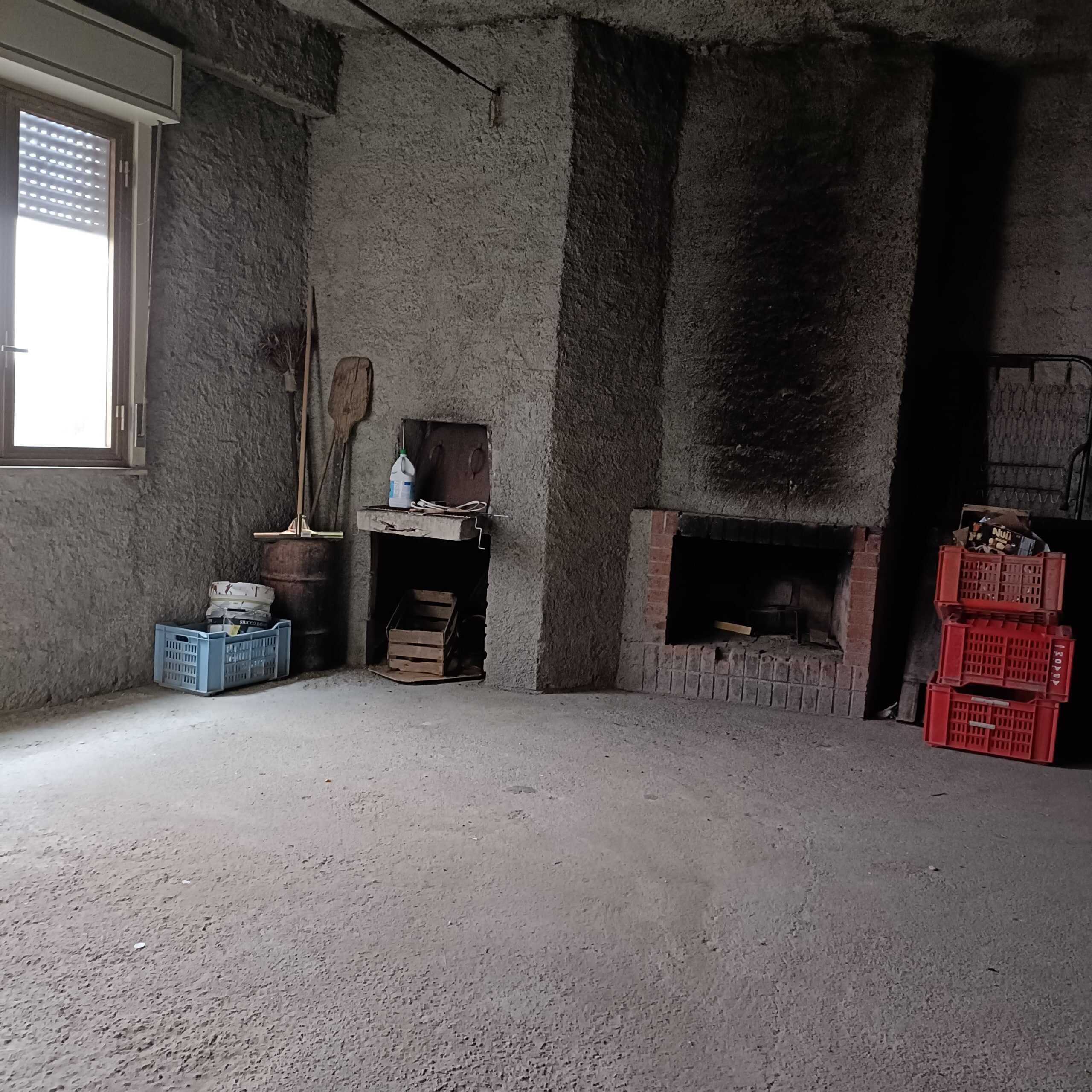 Stanza interna con camino in pietra, pareti in muratura grezza e pavimento in cemento, con scatole di plastica rosse e blu, utensili da lavoro e finestra che lascia entrare luce naturale, ambiente rustico di una casa in costruzione o ristrutturazione.