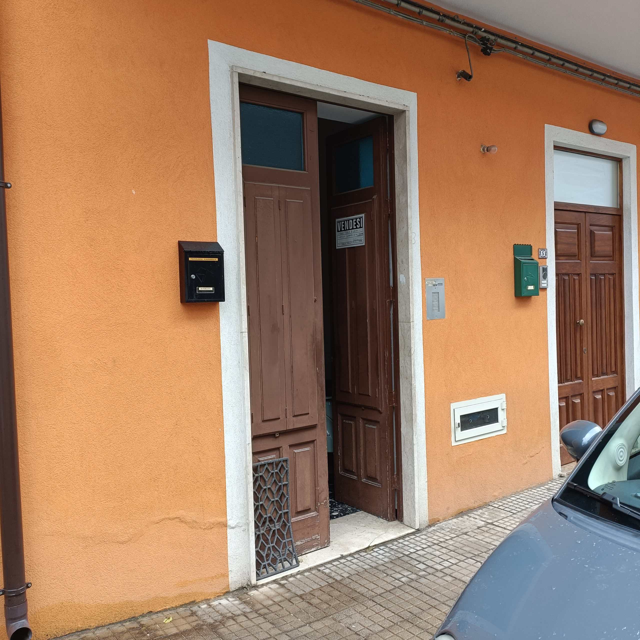 Porta di ingresso in legno con muro arancione e cassettina delle poste nera, con annuncio di vendita e citofono, scena in strada con pavimentazione e parte di vettura in primo piano.