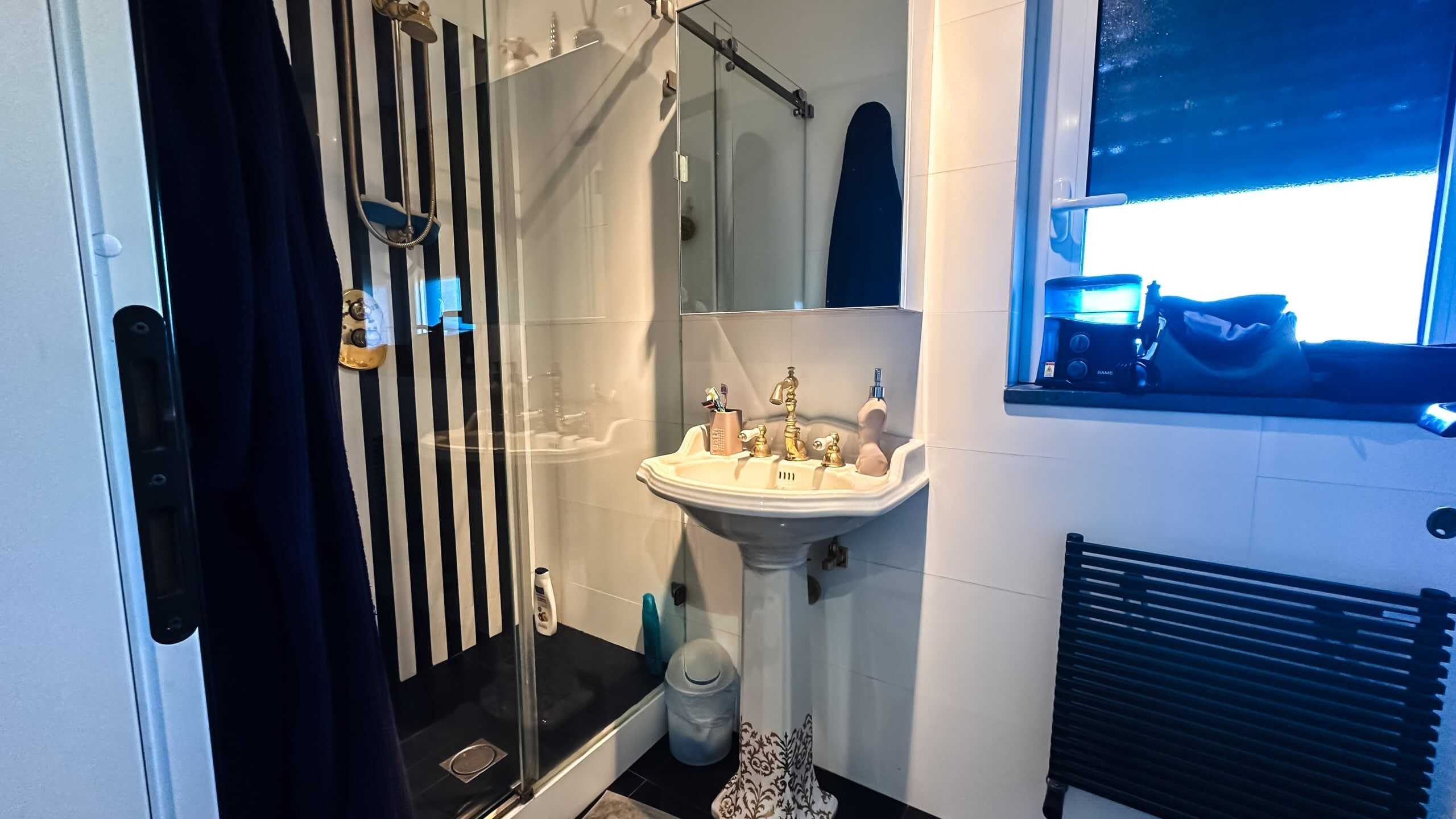 Doccia walk-in con cabina in vetro e piastrelle nere e bianche, lavabo antico con accessori e specchio, finestra con vista esterna, dettagli di arredo bagno eleganti e funzionali.