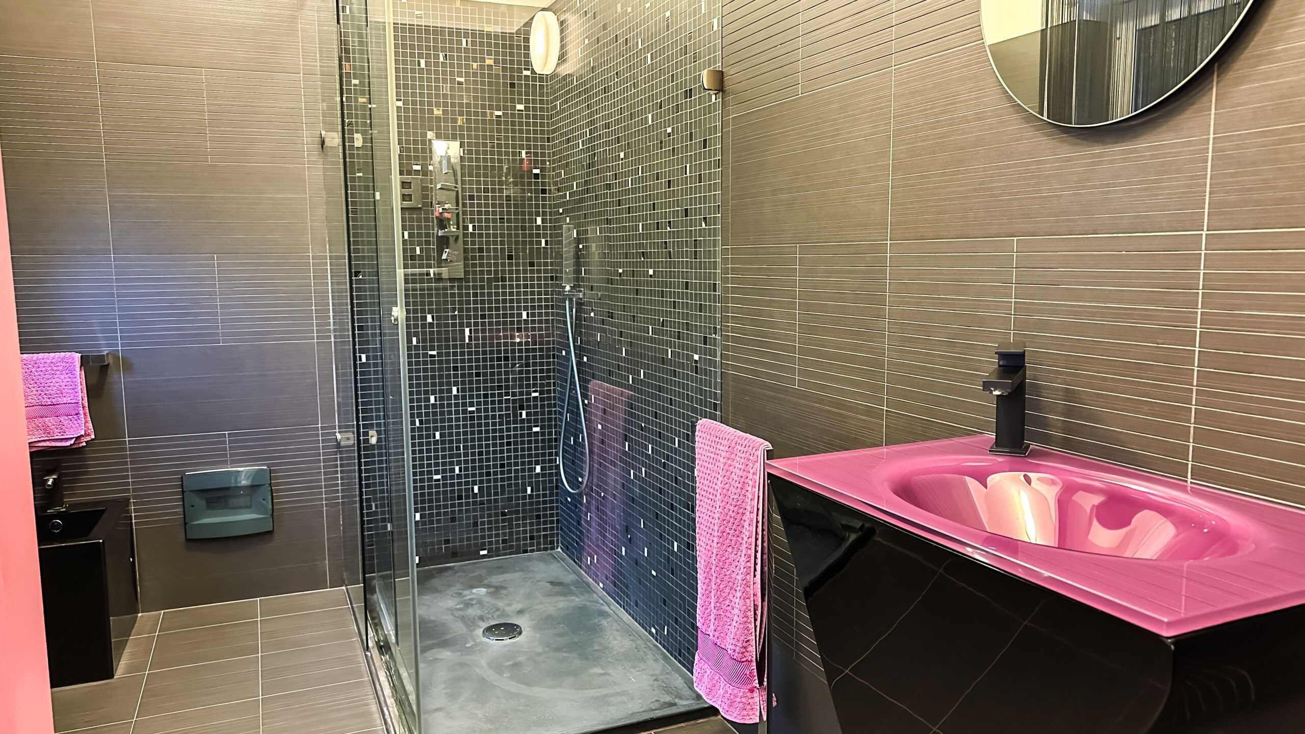 Lavabo rosa in bagno moderno con piastrelle marroni e zona doccia in mosaico nero, completo di accessori e specchio rotondo.