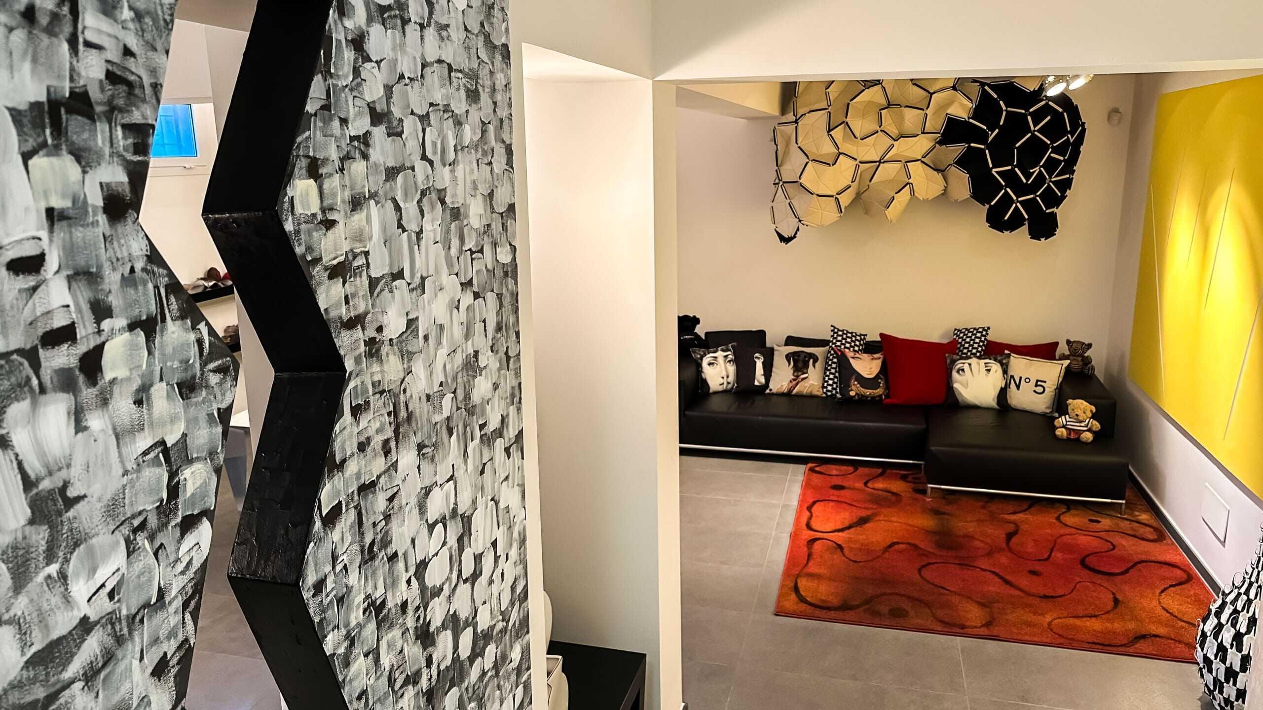 Colorato soggiorno con divano nero decorato con cuscini artistici, tappeto arancione, pareti decorative con elementi geometrici e moderni, ambientazione accogliente e stilosa per la casa.