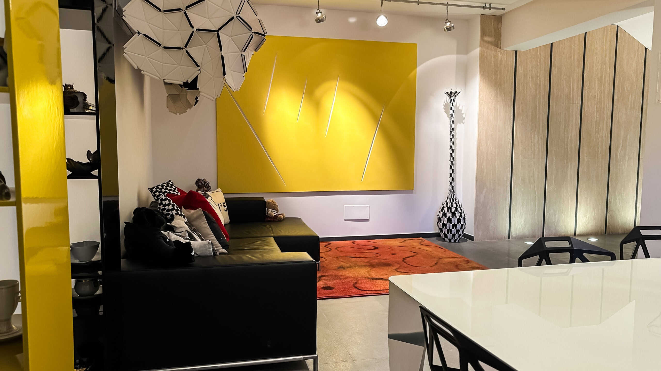 Soggiorno moderno con parete gialla, divano nero, decorazioni colorate e luce d'accento, ambiente elegante e di design.