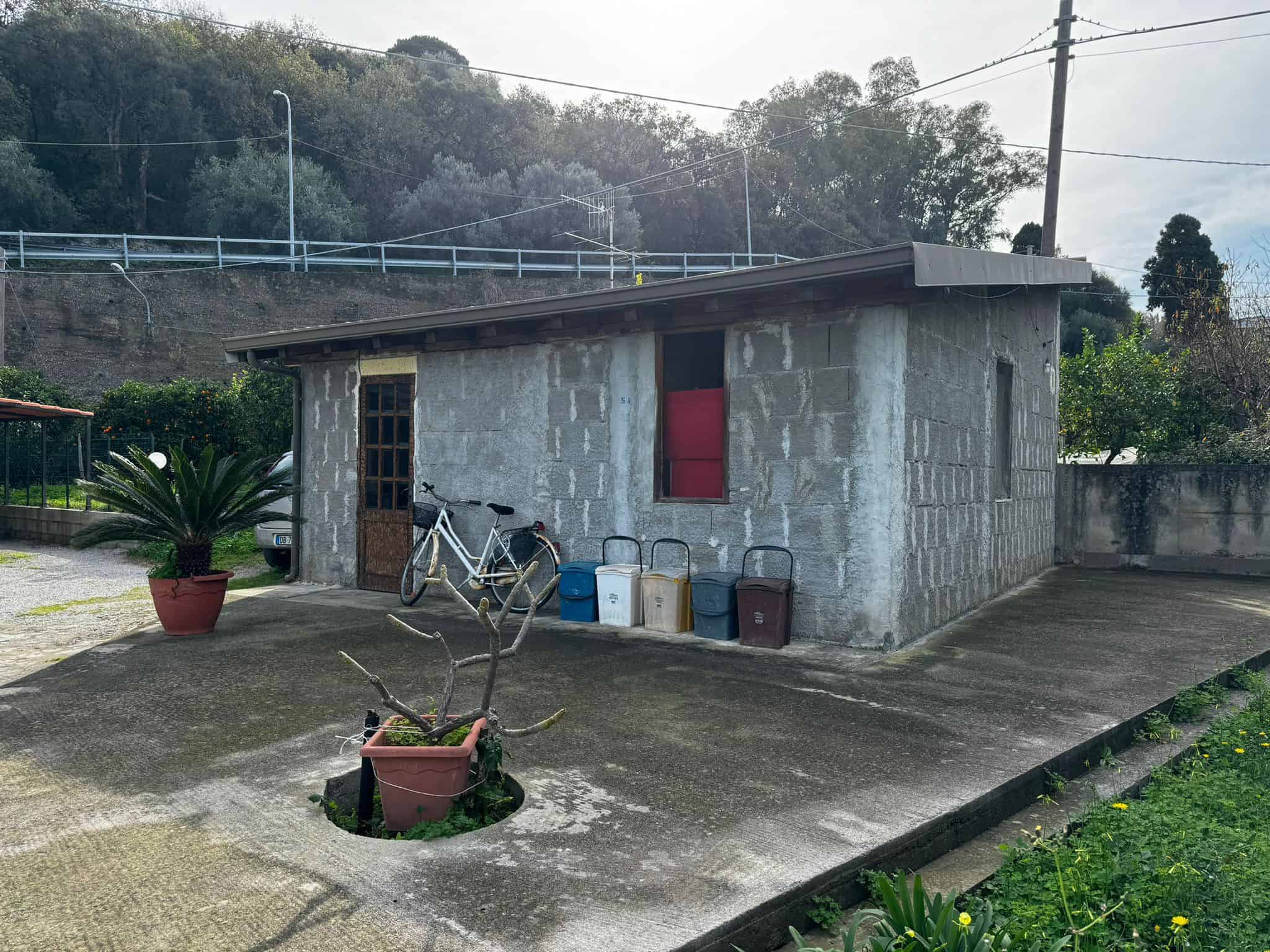 Vecchio magazzino in cemento a un piano con bici, contenitori e piante, situato in un contesto rurale o di campagna. Casa di campagna o struttura di deposito, prettamente funzionale, con pareti in pietra e copertura in lamiera.