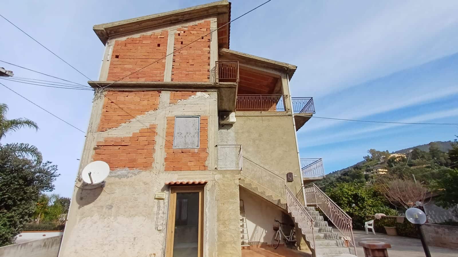 Vecchia casa ristrutturata con balcone e scale esterne, situata in zona residenziale di montagna con vista panoramica. Edificio in stile tradizionale italiano, ideale per investimenti immobiliari.
