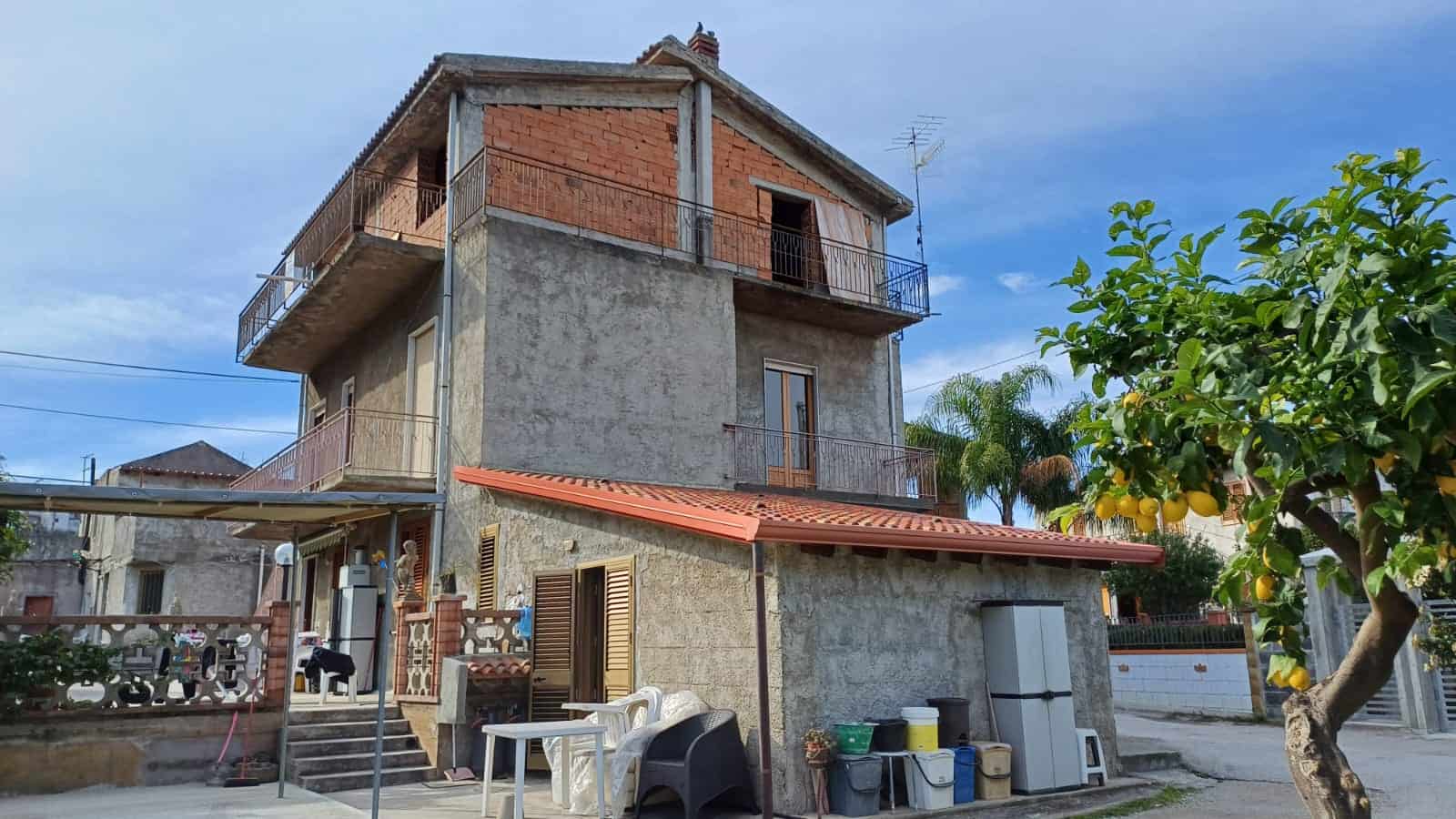 Vecchia casa in pietra a tre piani con balconi e terrazza, circondata da alberi di limoni in un ambiente residenziale, perfetta per investimenti immobiliari e ristrutturazioni.