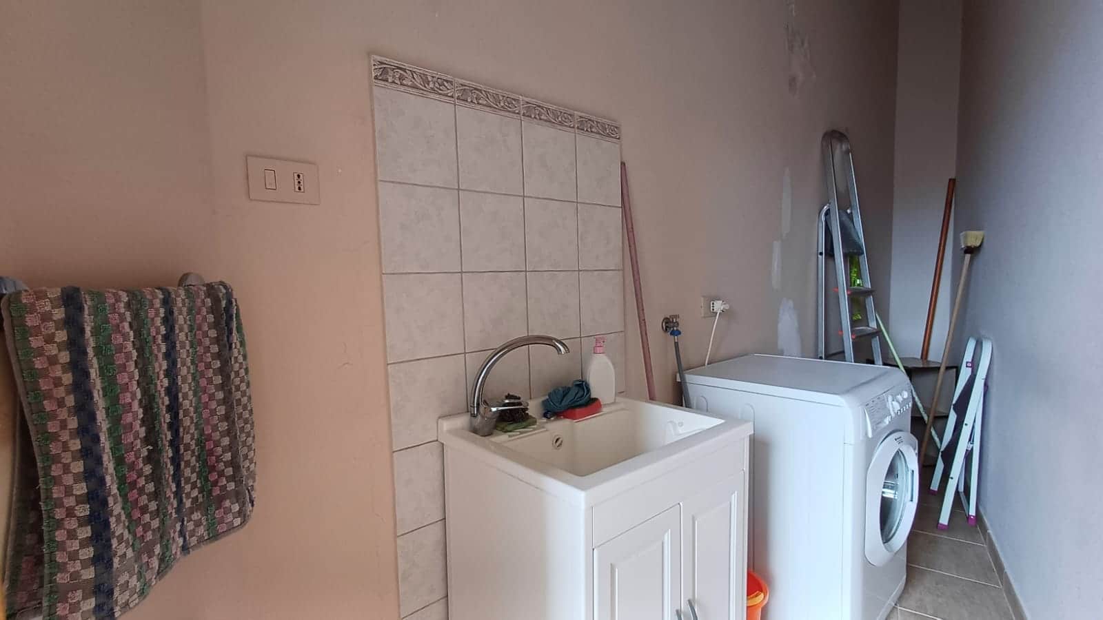 Lavanderia domestica con lavatrice e lavandino, scaffale e mestoli, ideale per servizi di pulizia e gestione della casa, in ambiente funzionale e pratico.