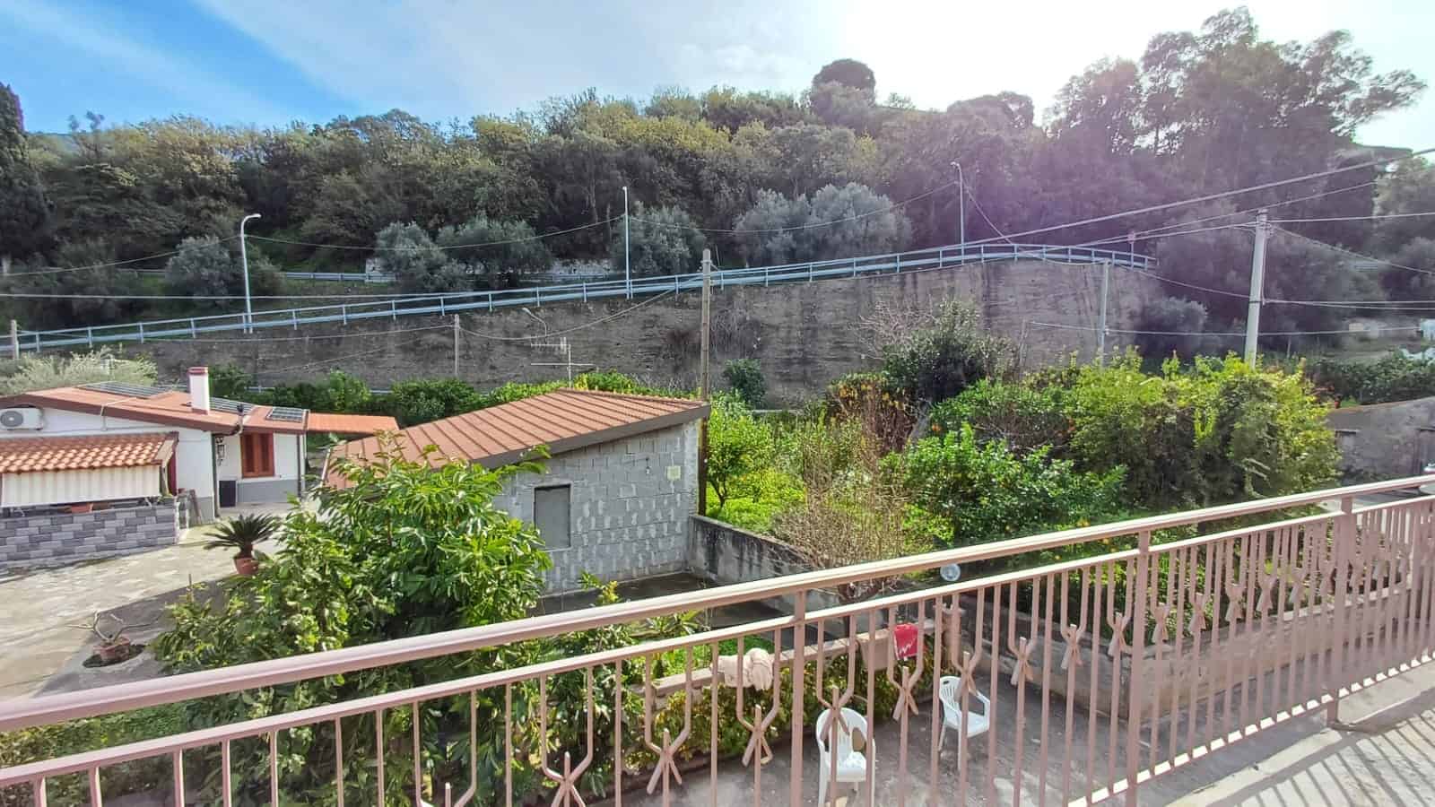 Vista panoramica su un giardino privato con piante e alberi, di fronte a una collina verdeggiante con case e linee elettriche, ideale per immobili immersi nella natura.