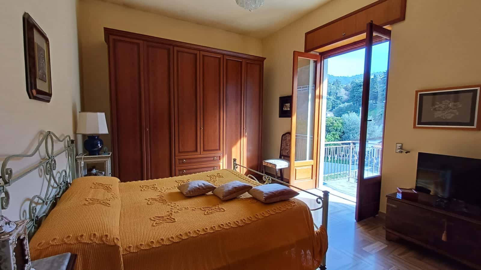 Arredo camera da letto con balcone vista mare, armadio in legno, letti con copriletto arancione, televisore su mobile, ambienti accoglienti e luminosi, stile classico e confortevole.