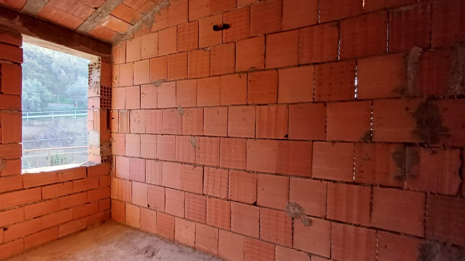 Finestra in muratura di mattoni rossi in una casa in fase di costruzione, vista verso l'esterno con elementi di sicurezza e paesaggio naturale.