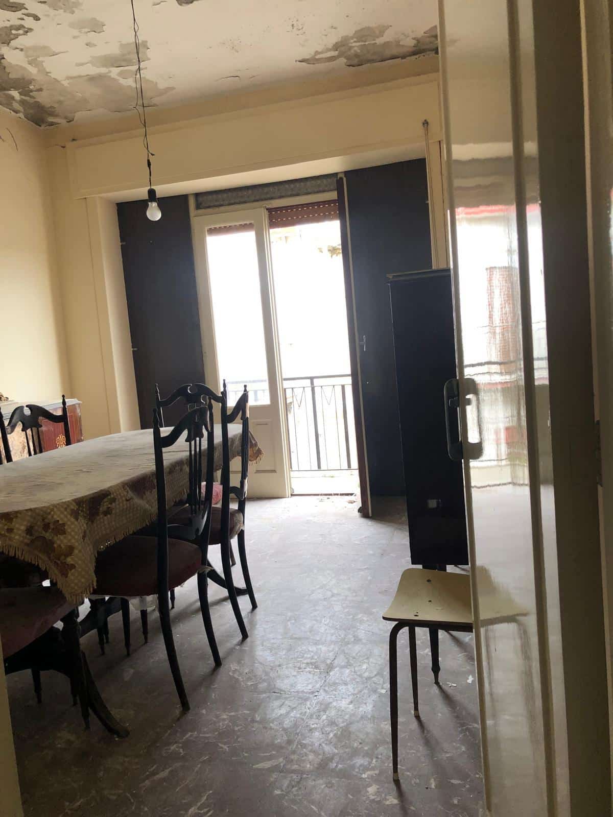 Soggiorno da ristrutturare con balcone, ingresso all'area living, pareti in fase di pittura, arredo datato, parquet usurato, ambiente abitabile, luce naturale.