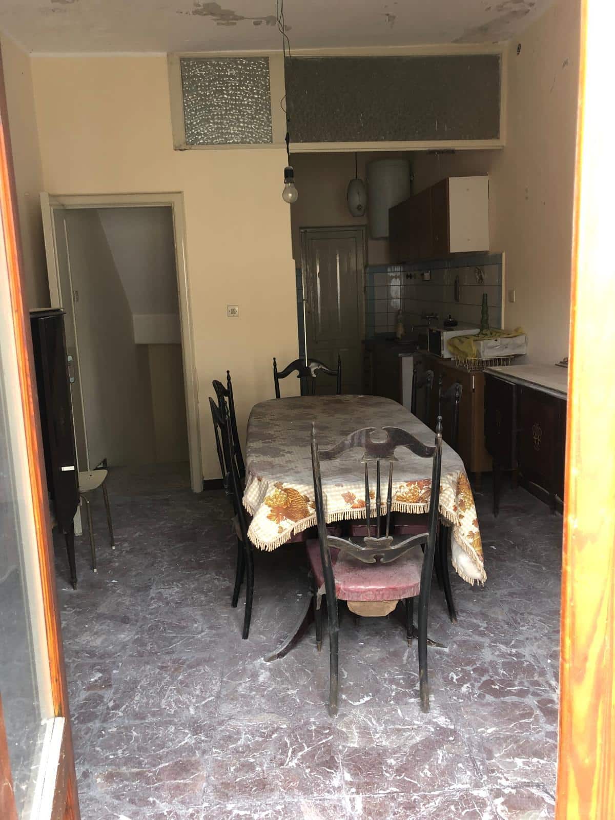 Vecchia cucina abitabile con tavolo rotto, sedie usurate e cucina vecchio stile, ambientazione di immobile da ristrutturare, vista da una porta aperta verso l’interno della casa, pavimento in marmo logoro, soluzione abitativa in vendita o affitto.