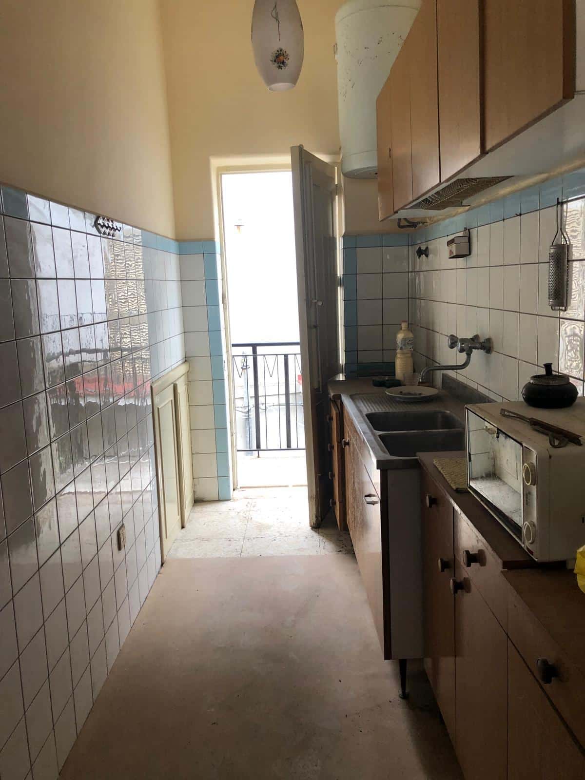 Finestra con balcone che dà su un appartamento, cucina con lavello, microonde, armadietti in legno e pareti rivestite con piastrelle bianche e azzurre, condizione abitativa da ristrutturare.