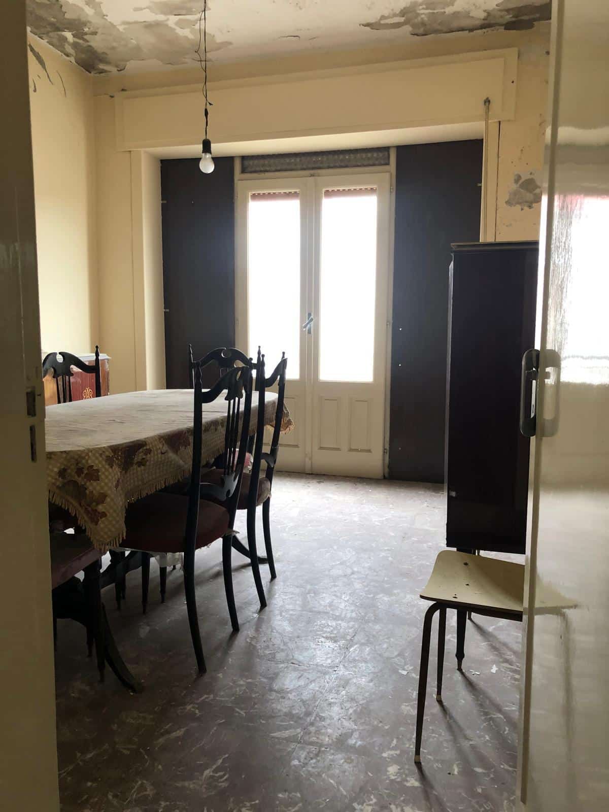 Appartamento da ristrutturare con sala da pranzo, grande finestra e pareti in stile classico, ideale per investimenti immobiliari o rinnovamento.