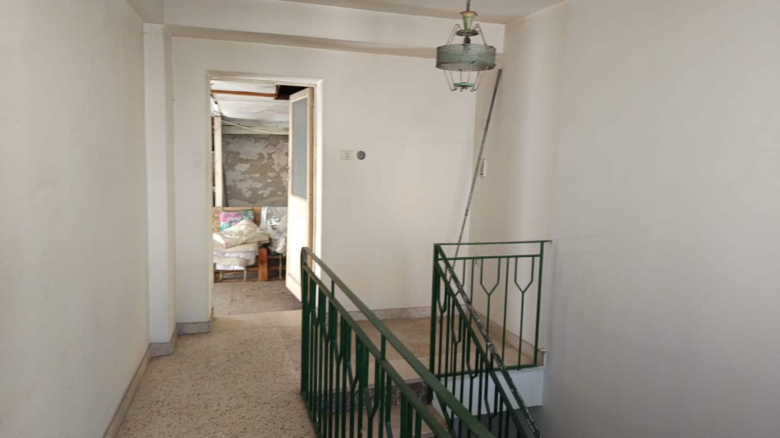 Vano scala e corridoio in un appartamento da ristrutturare con pareti bianche e pavimento in terrazzo, vista verso una stanza con oggetti e materiali di restauro.