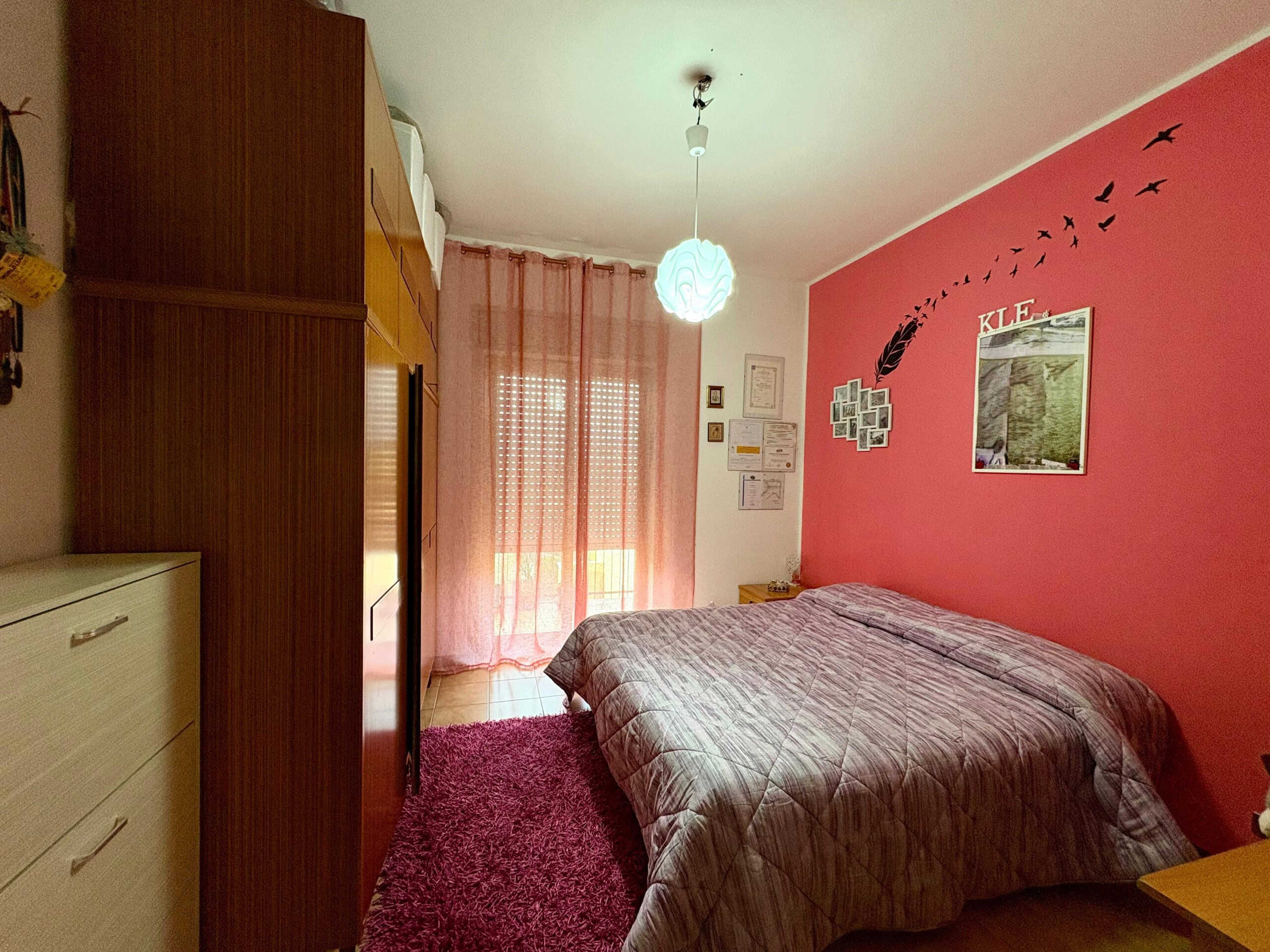 Camera da letto matrimoniale con pareti rosa, arredi in legno, comodo letto, specchio grande, armadio capiente, decorazioni sulle pareti, tende leggere, pavimento in ceramica, luminosa e accogliente, ideale per affitto o acquisto immobiliare.