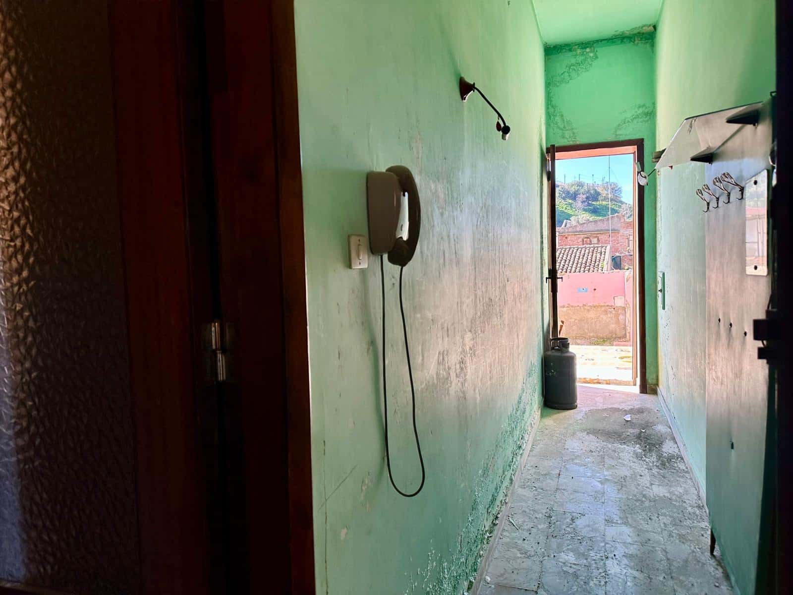 Telefono a filo muro verde in corridoio con vista all'esterno, pareti e pavimento usurati, luce naturale, ambiente essenziale e rustico.