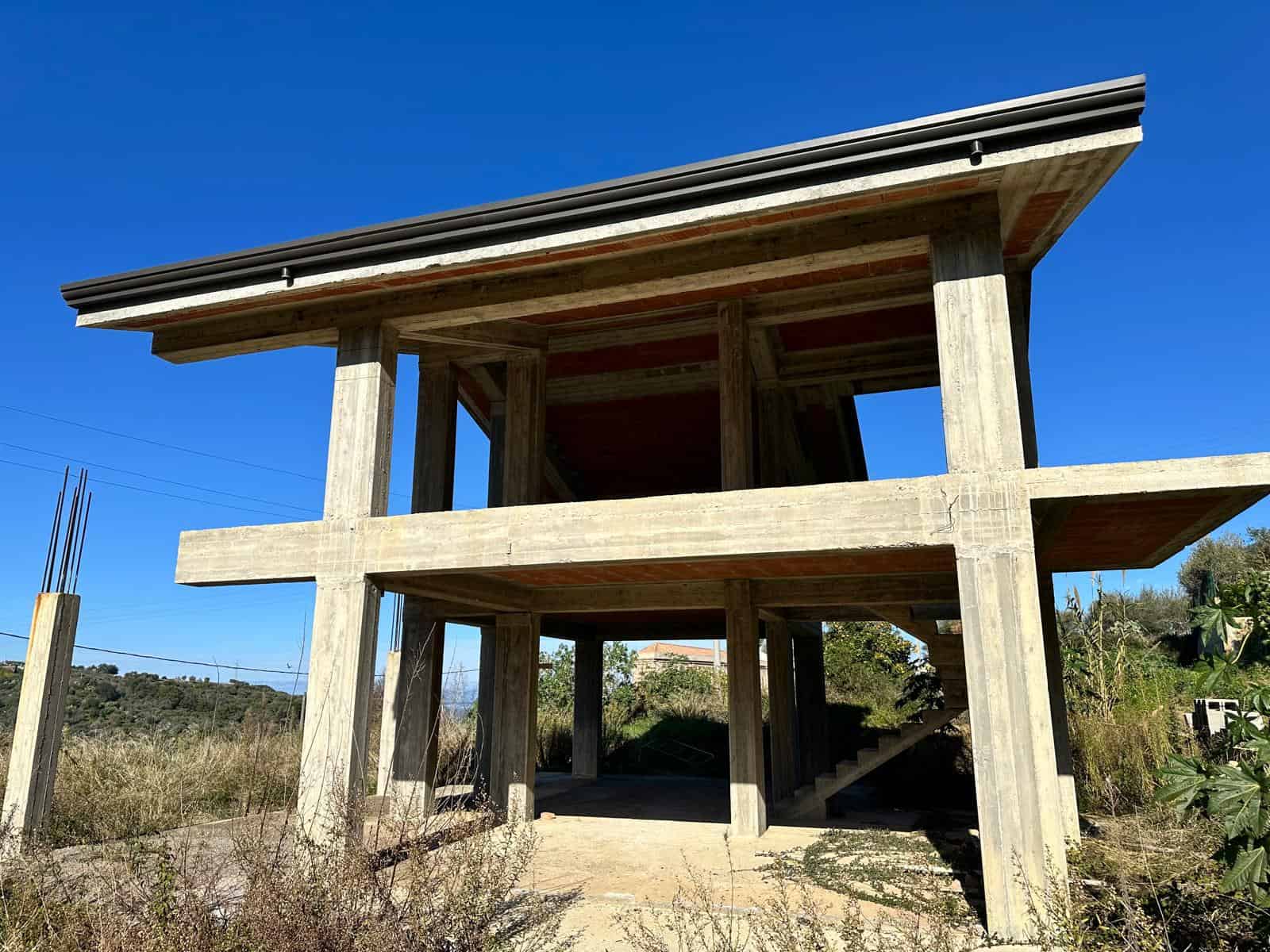 Cantiere di costruzione di una casa moderna in cemento armato con struttura a due piani e vista su un paesaggio naturale, sotto un cielo azzurro.