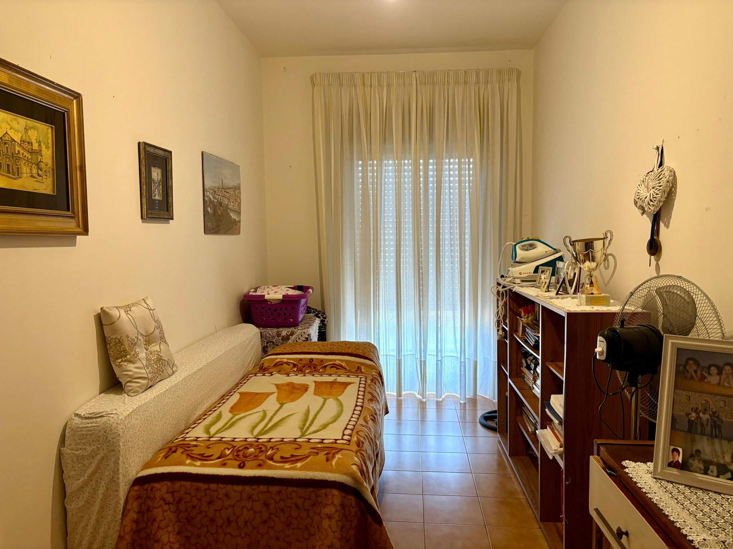Voglio un'anteprima di una stanza da letto con arredi semplici, decorazioni di famiglia, quadri appesi alle pareti e una finestra con tende leggere, creando un'atmosfera accogliente e familiare.