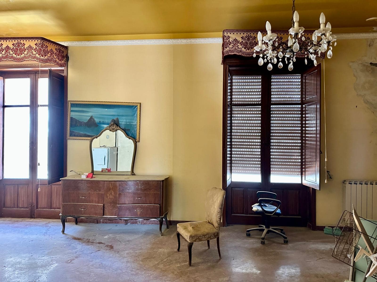 Antico soggiorno con grandi finestre in legno, mobile in stile vintage con specchio, sedie in velluto e lampadario in cristallo, ambientazione storica e spaziosa.