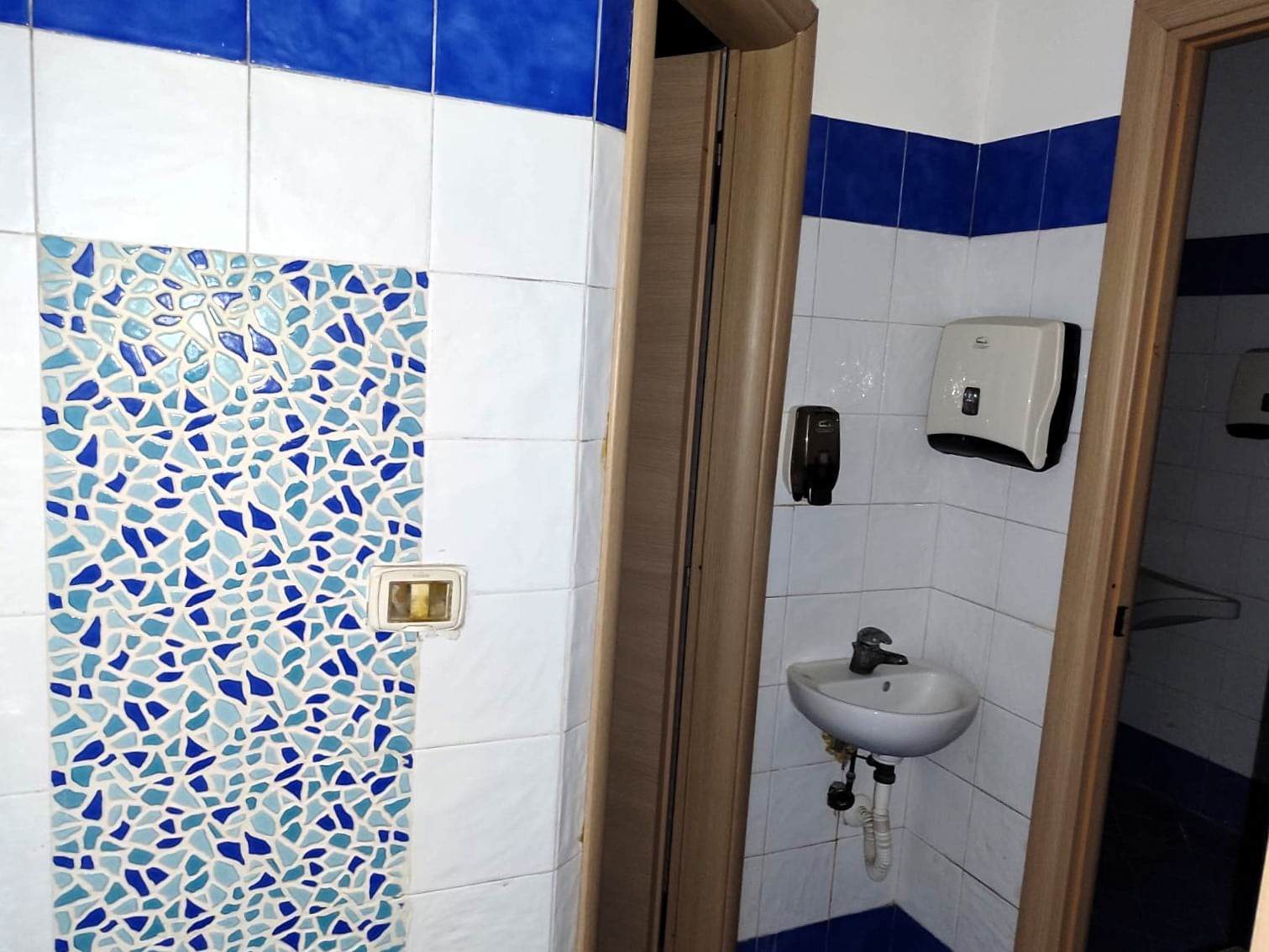 Lavabo e muri del bagno con piastrelle bianche e decorazioni blu, sanitari e dispenser di sapone in un ambiente pulito e funzionale.