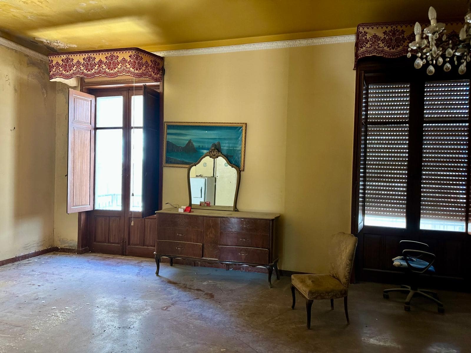 Vecchio soggiorno di casa con mobili d'epoca, finestra con tende a stucchi in legno, pareti gialle, soffitto con decorazioni e lampadario, ambiente da ristrutturare, stile vintage, spazio interno ampio.