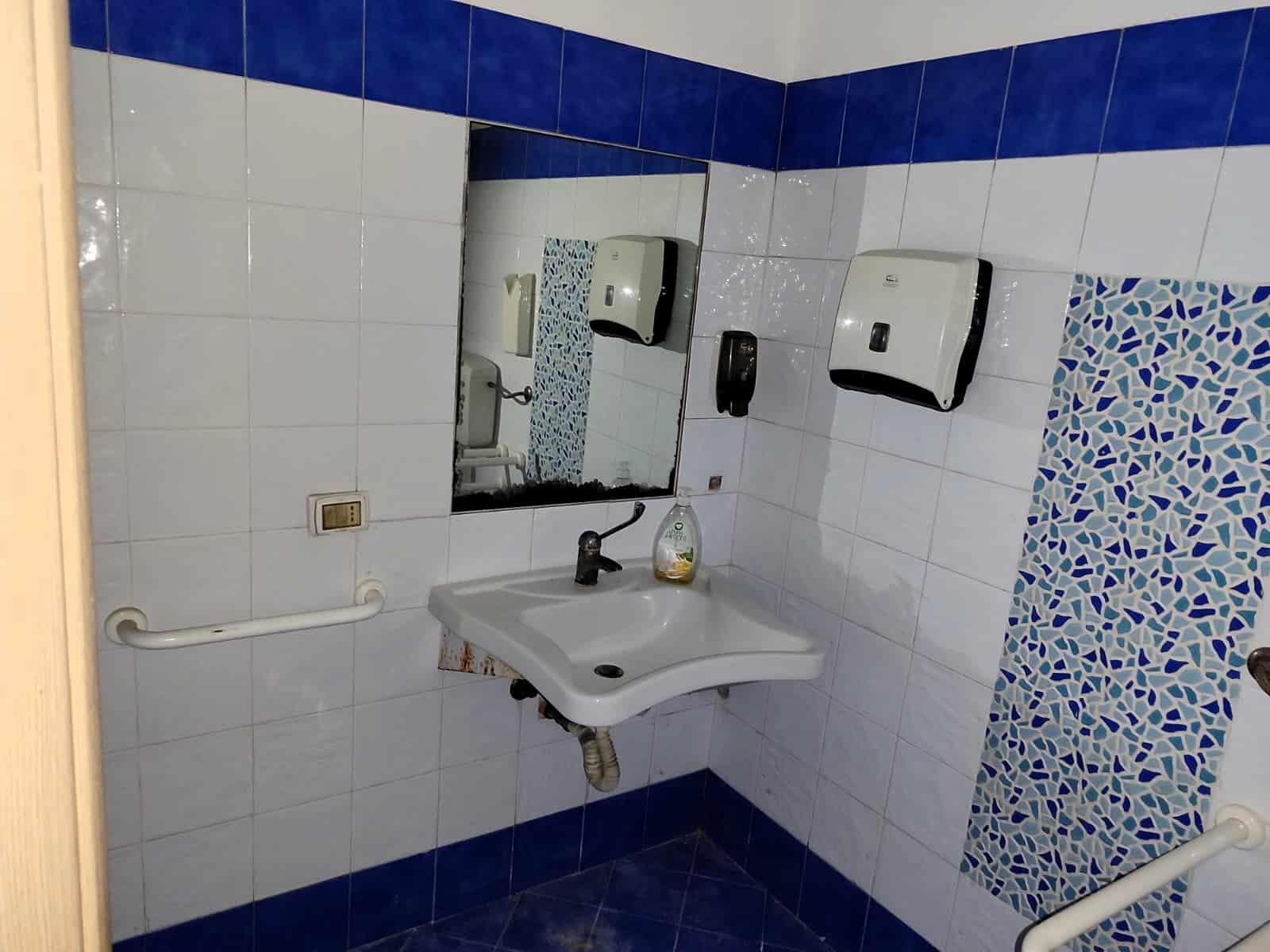 Lavabo e sanitari in bagno pubblico con piastrelle bianche e dettagli blu, specchio fronte al lavabo, dispenser sapone e asciugamani, ambiente pulito e funzionale per servizi igienici condivisi.