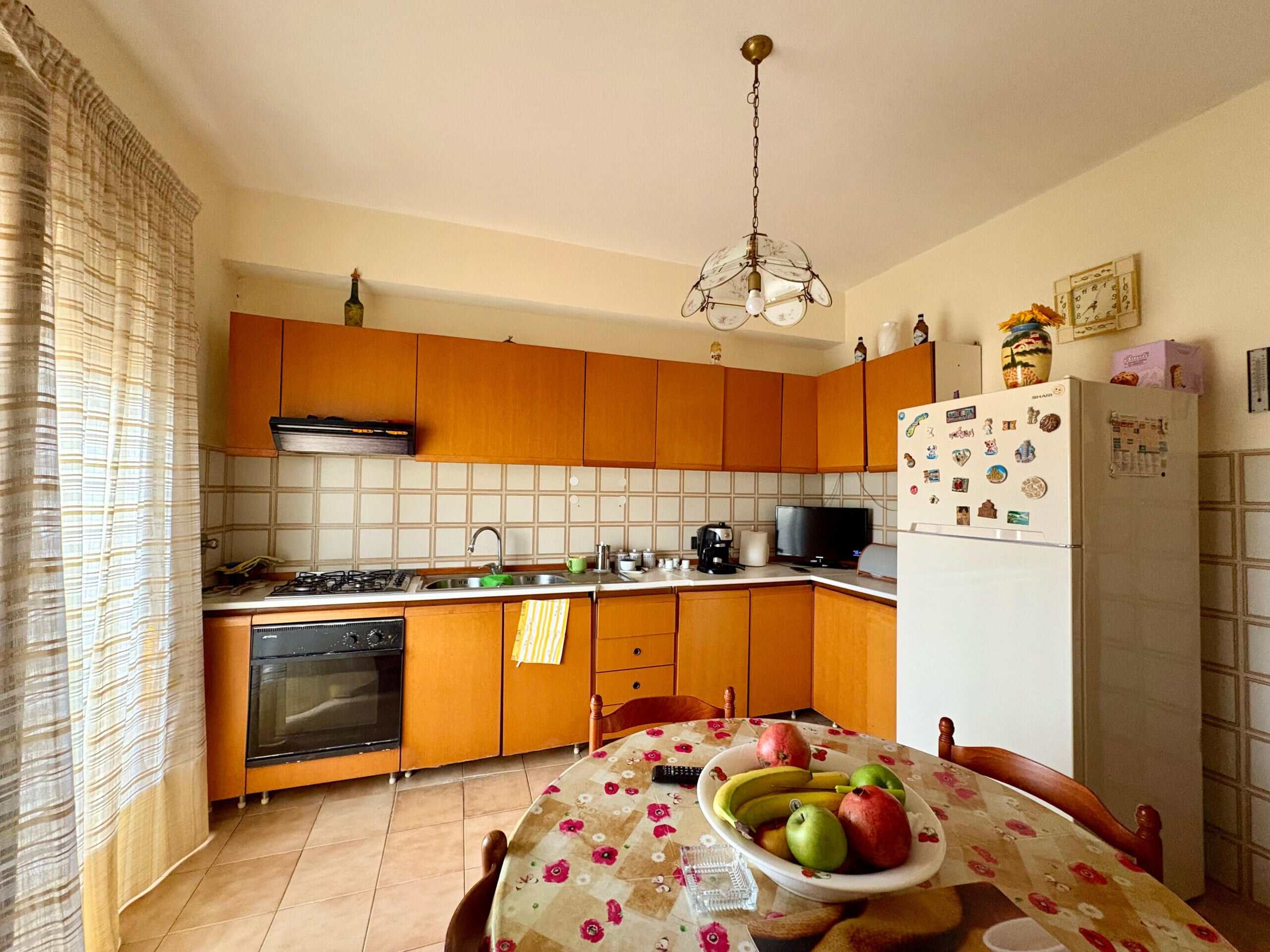 Finestra con tende leggere che illumina una cucina abitabile con mobili in legno, frigo, forno e tavolo con frutta fresca e tovaglia colorata, ambiente accogliente e funzionale.