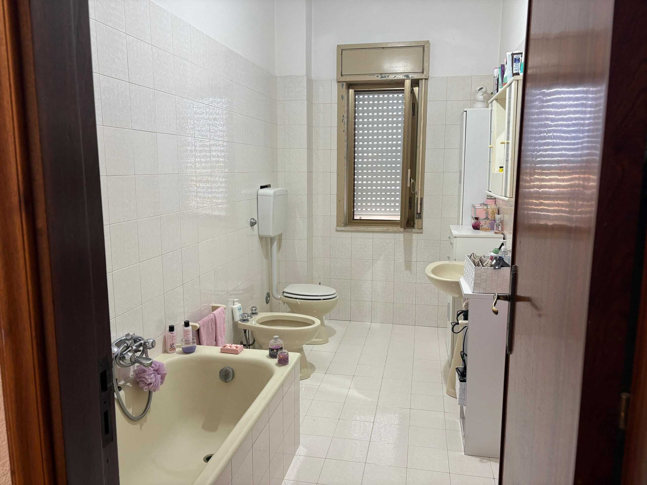 Bagno cucina in stile classico con vasca, lavabo e servizi igienici, finestra con avvolgibile, arredi semplici, piastrelle bianche, dettagli pratici e funzionali, ideale per ristrutturazione o aggiornamento.