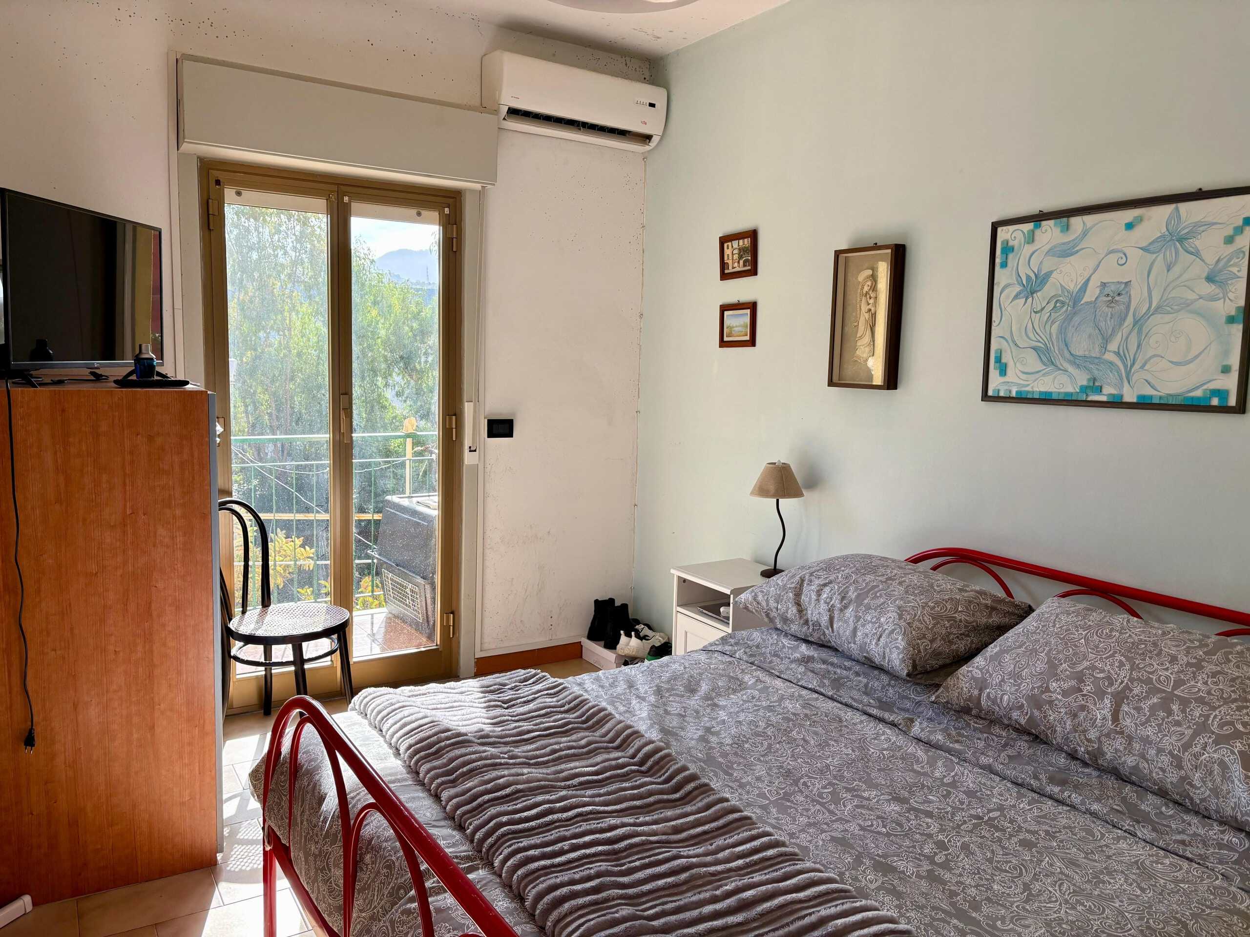 Balcone con vista su natura e montagne, camera da letto arredata con letto matrimoniale, quadri decorativi sulle pareti, aria condizionata, luce naturale, ambiente accogliente e tranquillo.