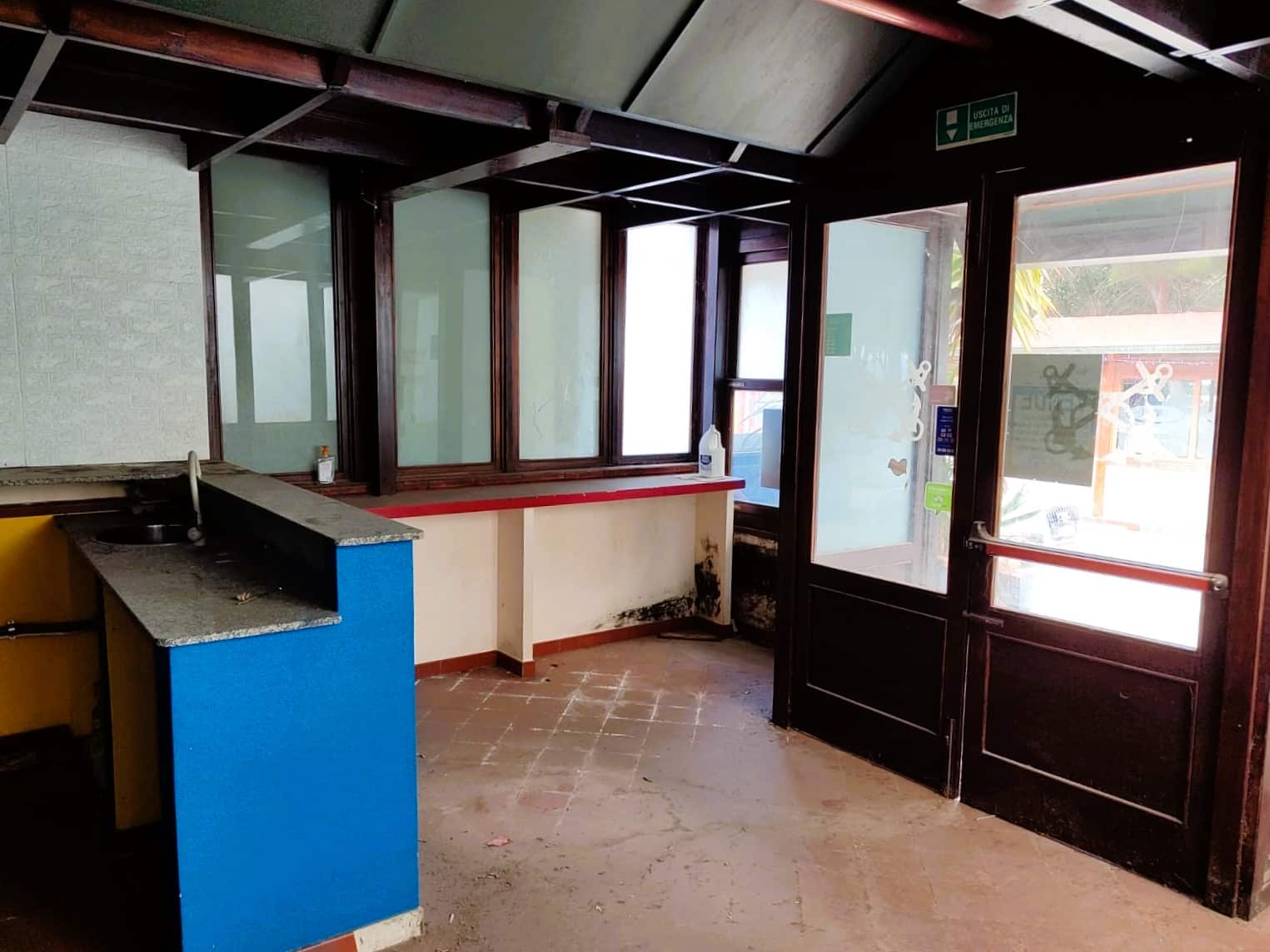 Ingresso di un locale commerciale con arredi datati, pareti in mattonelle, porta finestra e pannelli in vetro, ambientazione brutta e trascurata, poco luminoso e in condizioni di degrado.