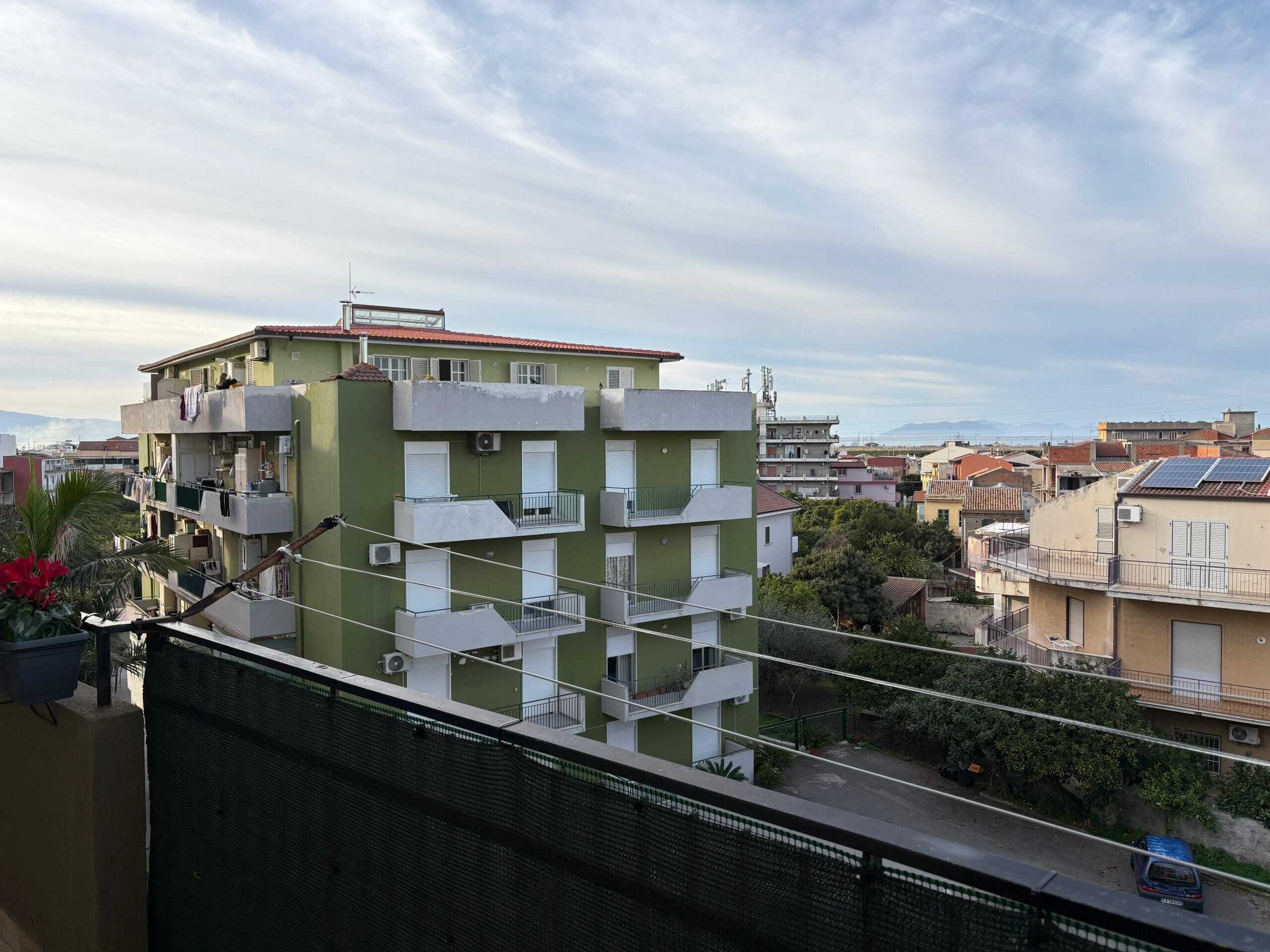 Balcone con vista su edifici residenziali e cielo nuvoloso, in zona urbana, con piante e linee di cavi elettrici.