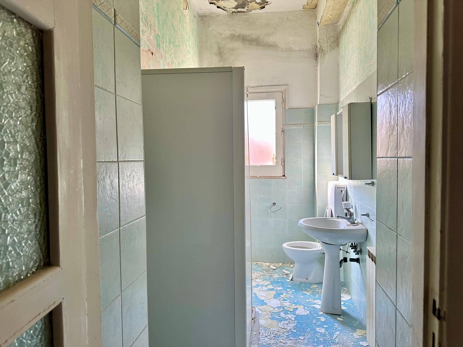Vecchio bagno in condizioni di abbandono, piastrelle stanche, pareti scolorite e soffitto rovinato, ambiente deteriorato senza arredi funzionanti, luce naturale che entra da una finestra sporca.