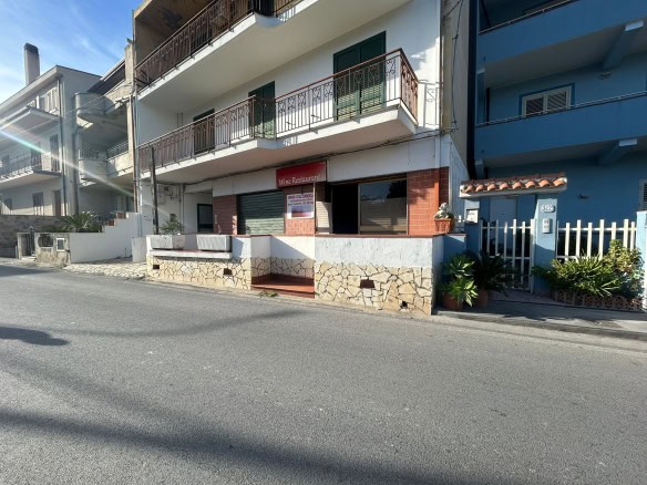 Negozio in affitto con progetto di ristrutturazione, pareti esterne di colore bianco, con piastrelle di pietra decorativa nel basamento, situato su strada trafficata a livello stradale, con ingresso pedonale e piante ornamentali, ideale per attività commerciali.