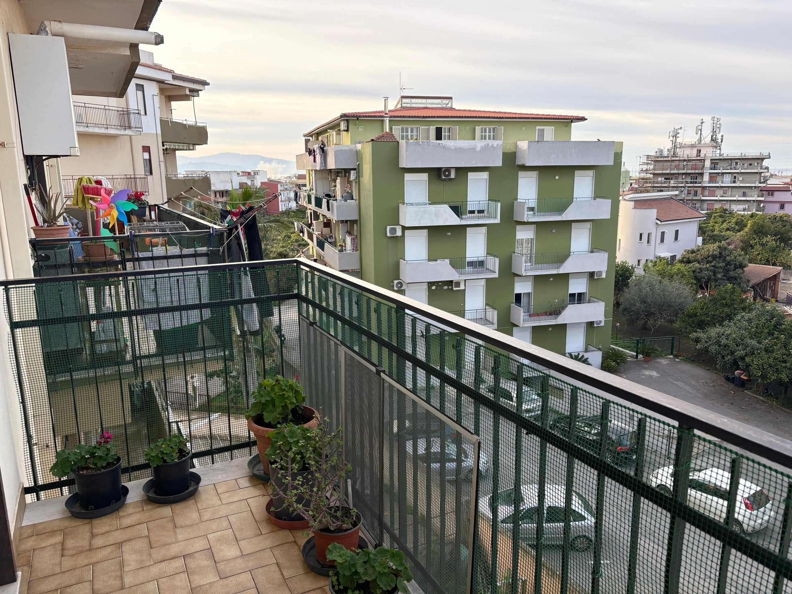 Balcone di un appartamento con piante in vaso e stendibiancheria, vista su edificio residenziale verde con balconi e sulle aree circostanti, in un contesto urbano con alberi e altri edifici.