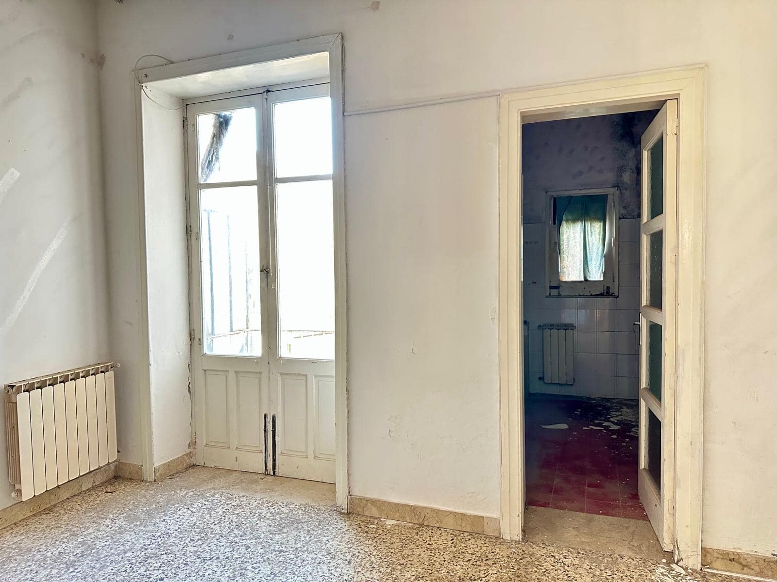 Scala di accesso luminosa e porta finestra in stile tradizionale, ambienti di casa da rinnovare, dettagli di abitazione in sofferenza, interni con pareti bianche e pavimento in marmo, vista su piccolo balcone, spazio da personalizzare, casa antica da ristrutturare, ambiente abitativo con caratteristiche storiche.